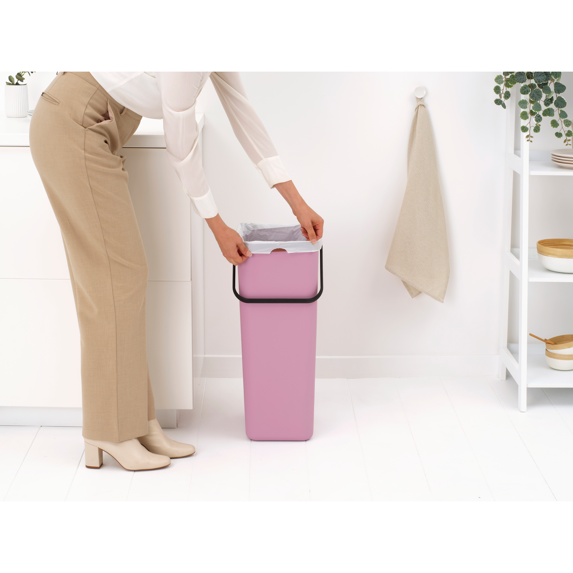Vrouw in beige broek en witte blouse plaatst vuilniszak in roze prullenbak.