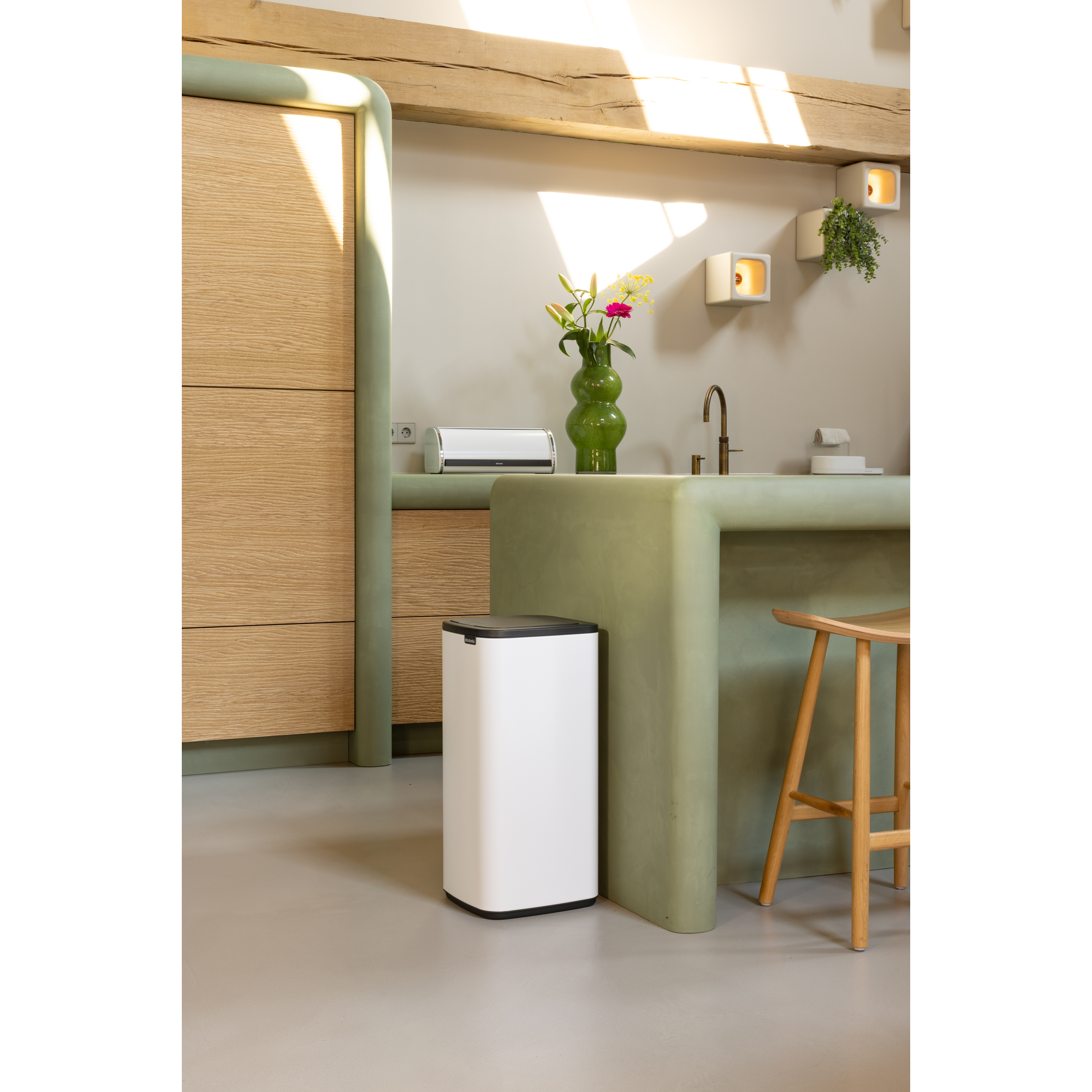 Moderne keuken met houten kasten, groen aanrecht, witte prullenbak en bloemen.