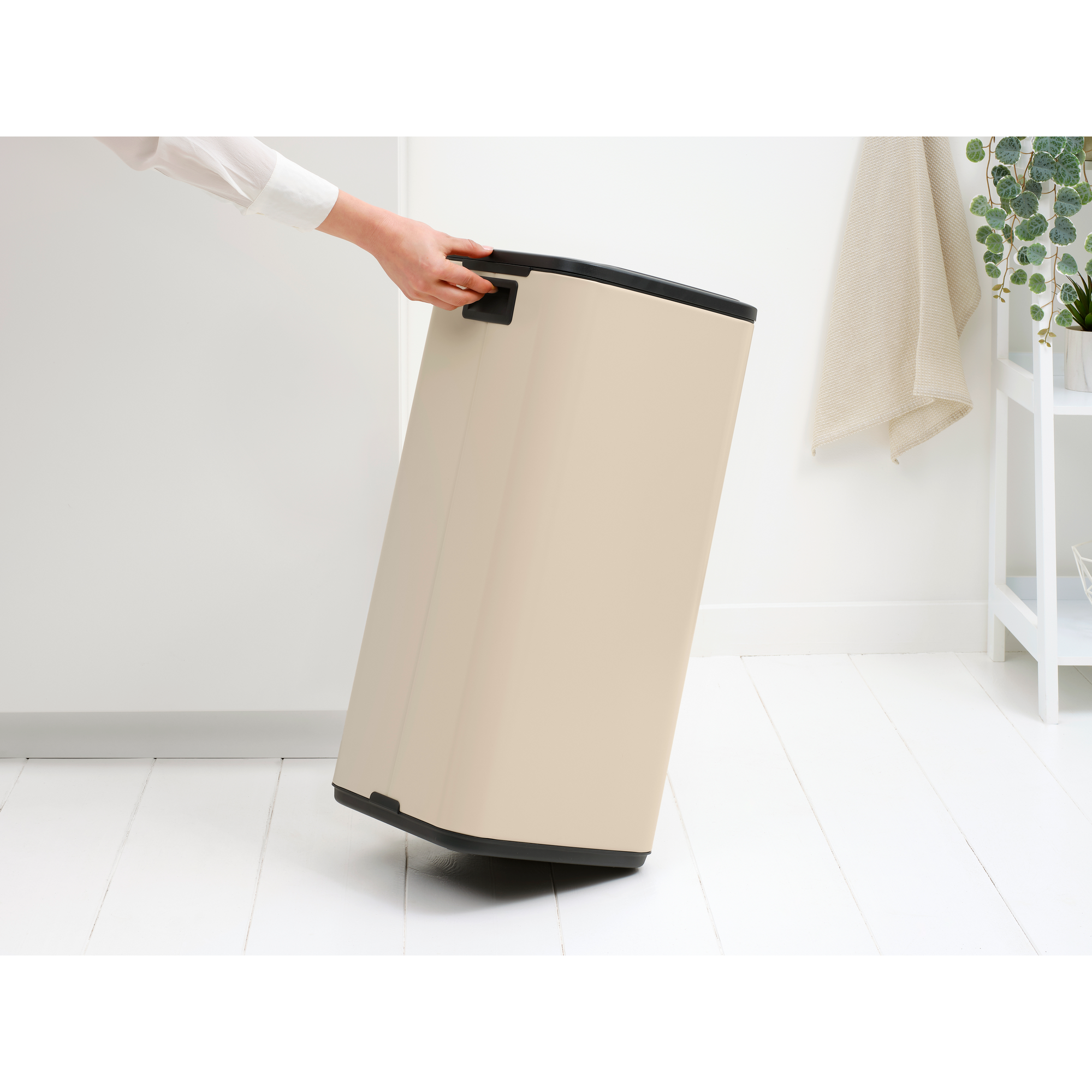 Een persoon in een wit overhemd houdt een beige wasmand met zwarte rand en handvatten vast, kantelend op een witte houten vloer.