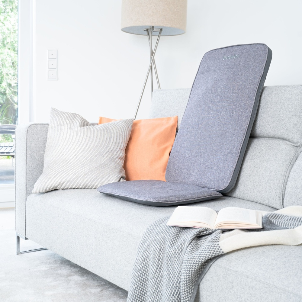 Szara sofa z poduszkami, masażerem, książką i kocem.