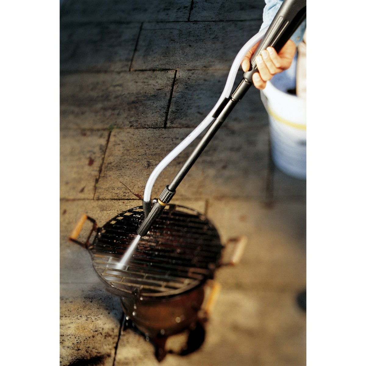 Una persona limpia una parrilla con una hidrolavadora sobre un camino pavimentado.