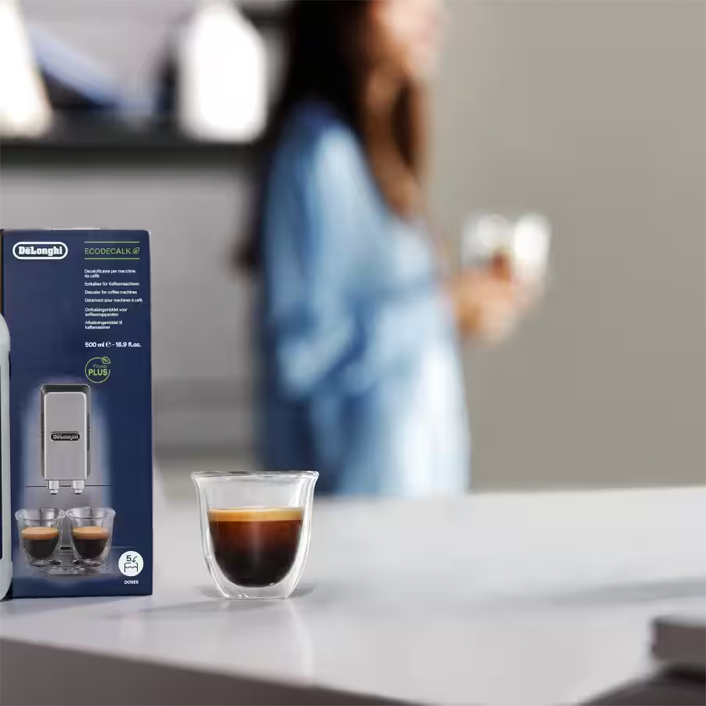 DeLonghi Entkalker-Verpackung neben einem Glas Espresso auf einem Tisch.