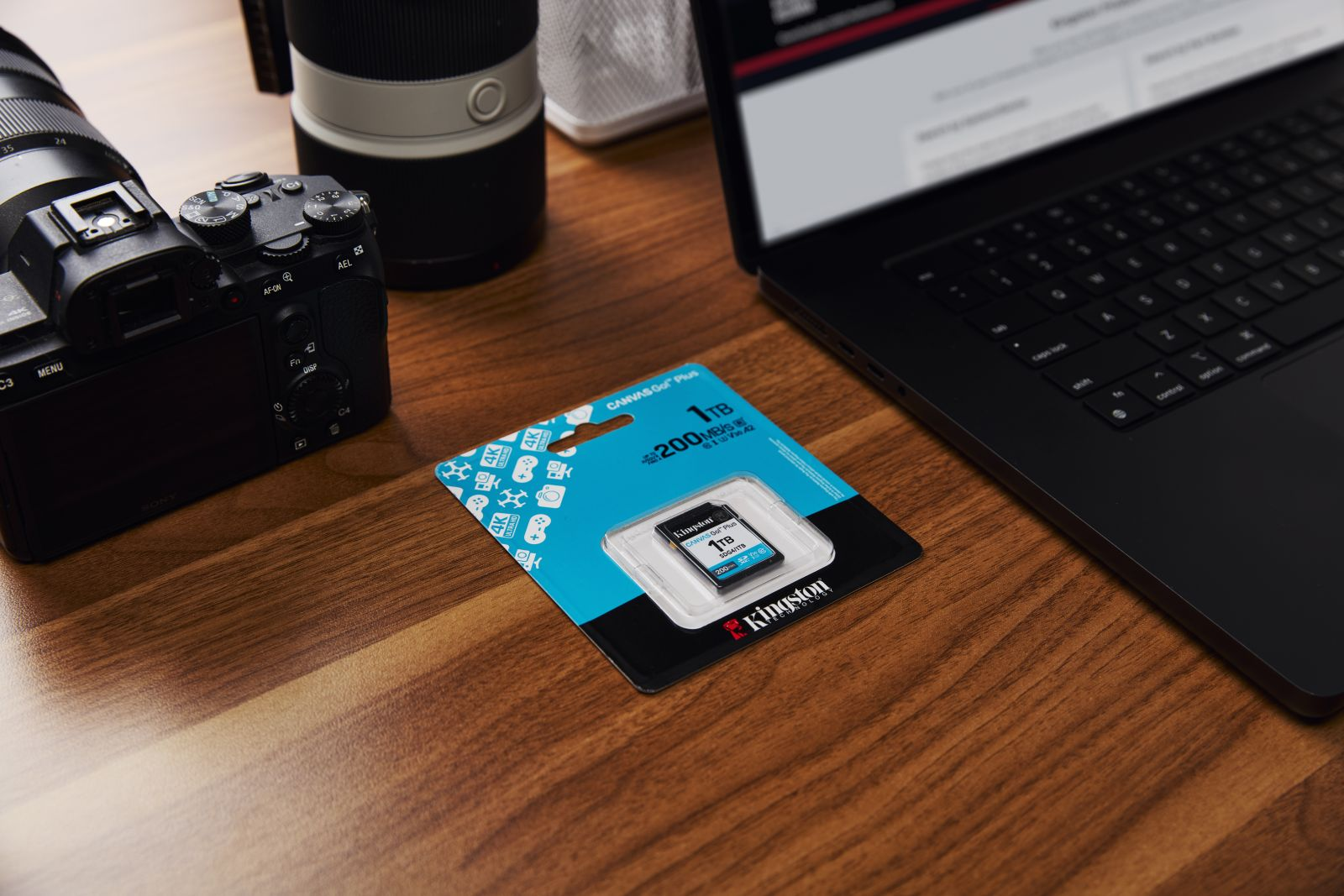1TB SD-kaart in blisterverpakking naast camera en laptop op houten tafel.