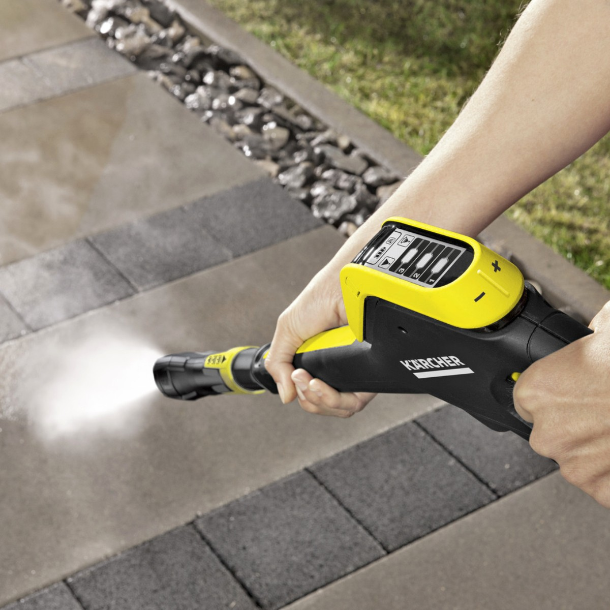 Una mano tiene un'idropulitrice Karcher gialla, spruzzando acqua su una superficie pavimentata.