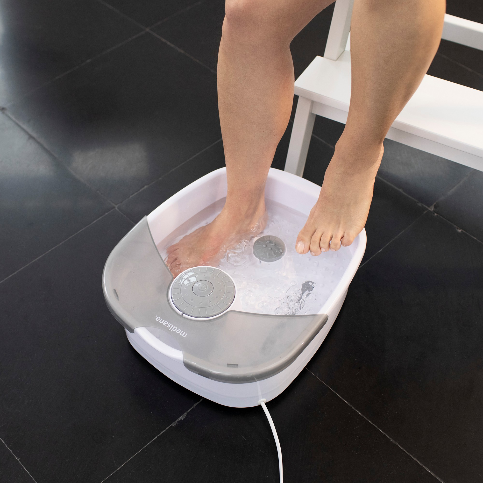 Die Füße einer Person tauchen in eine weiße Fußbadewanne mit Wasser. Der Boden ist schwarz gefliest.