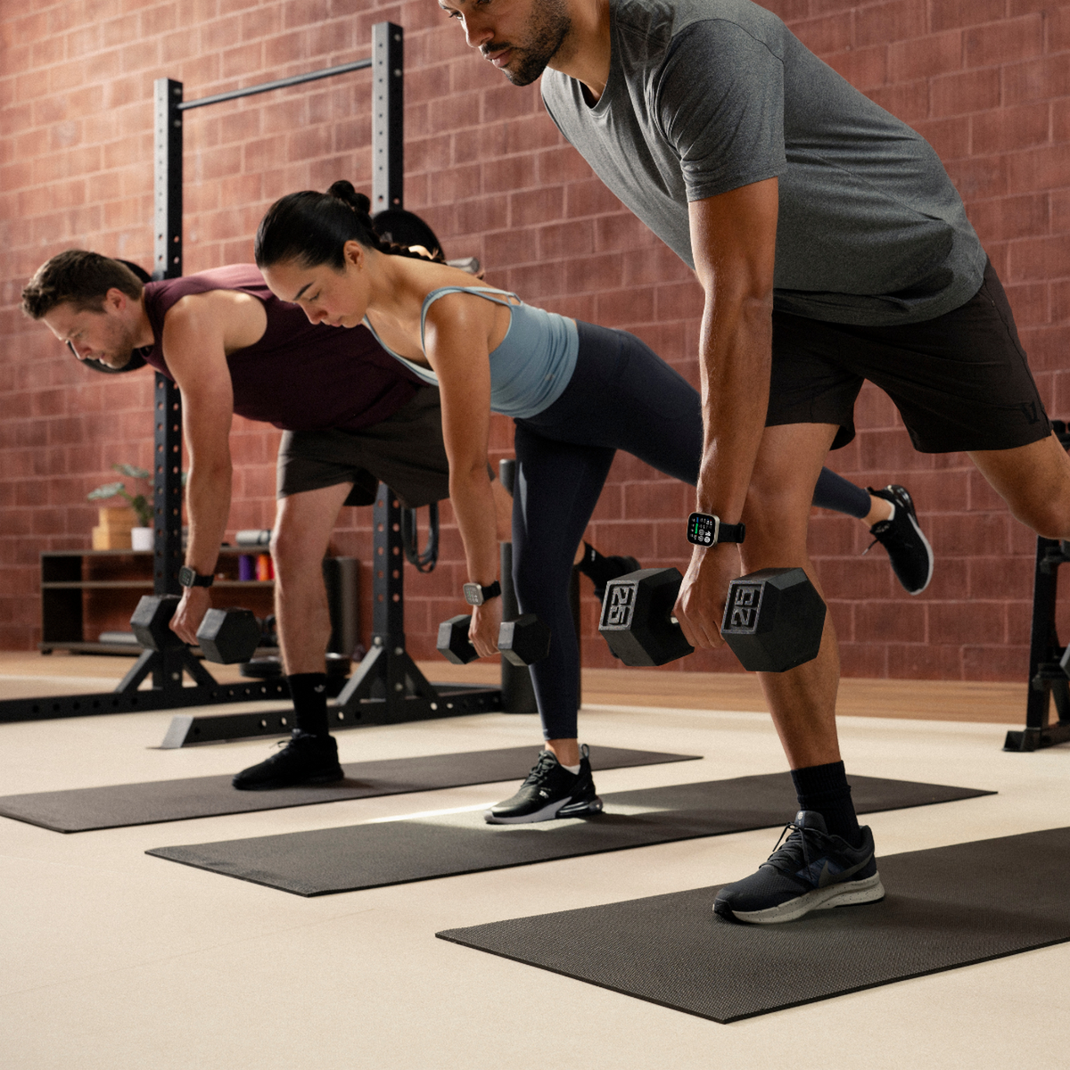 Tres personas hacen ejercicio con mancuernas en un gimnasio frente a una pared de ladrillos.
