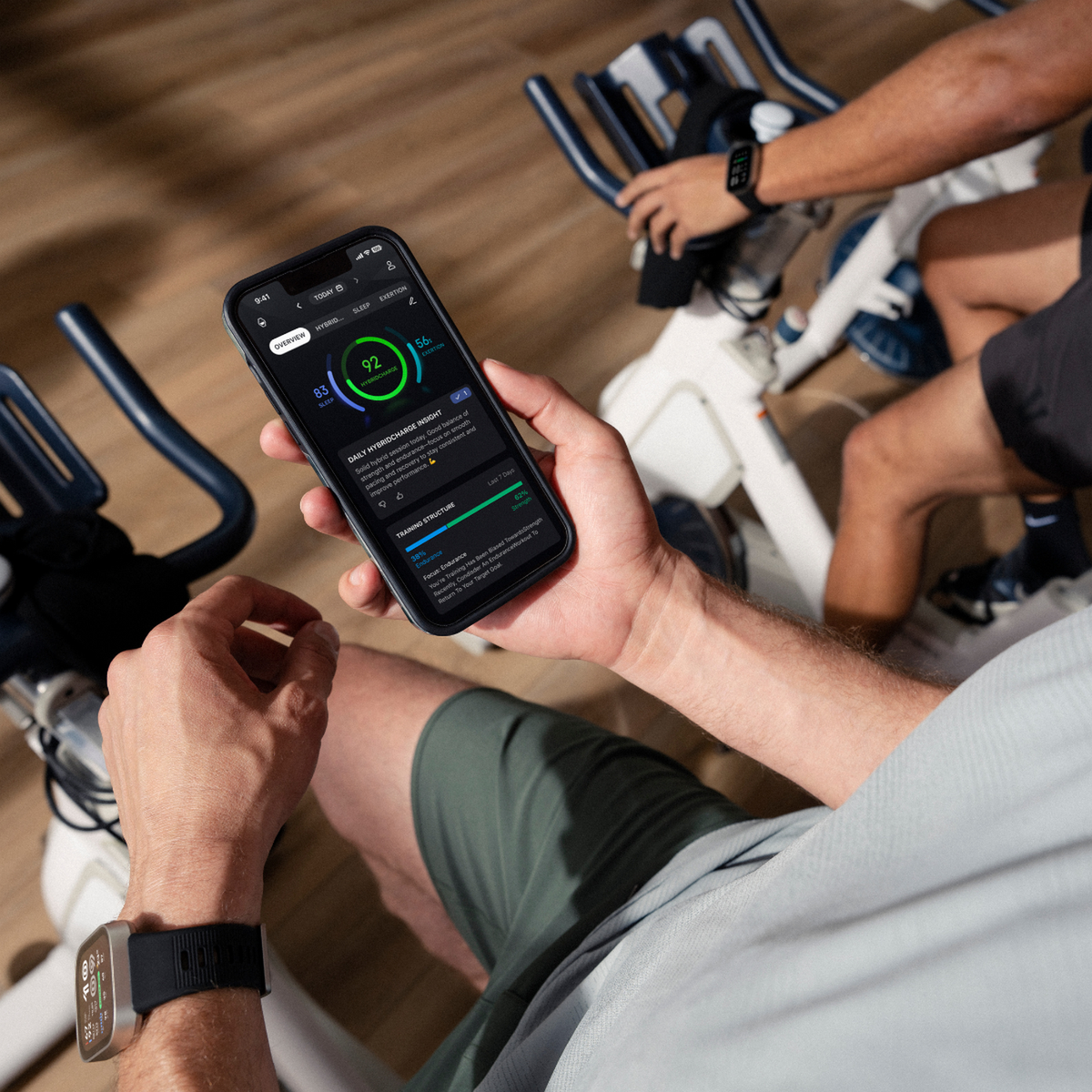 Hombre sostiene teléfono con app de fitness sentado en bicicleta estática.
