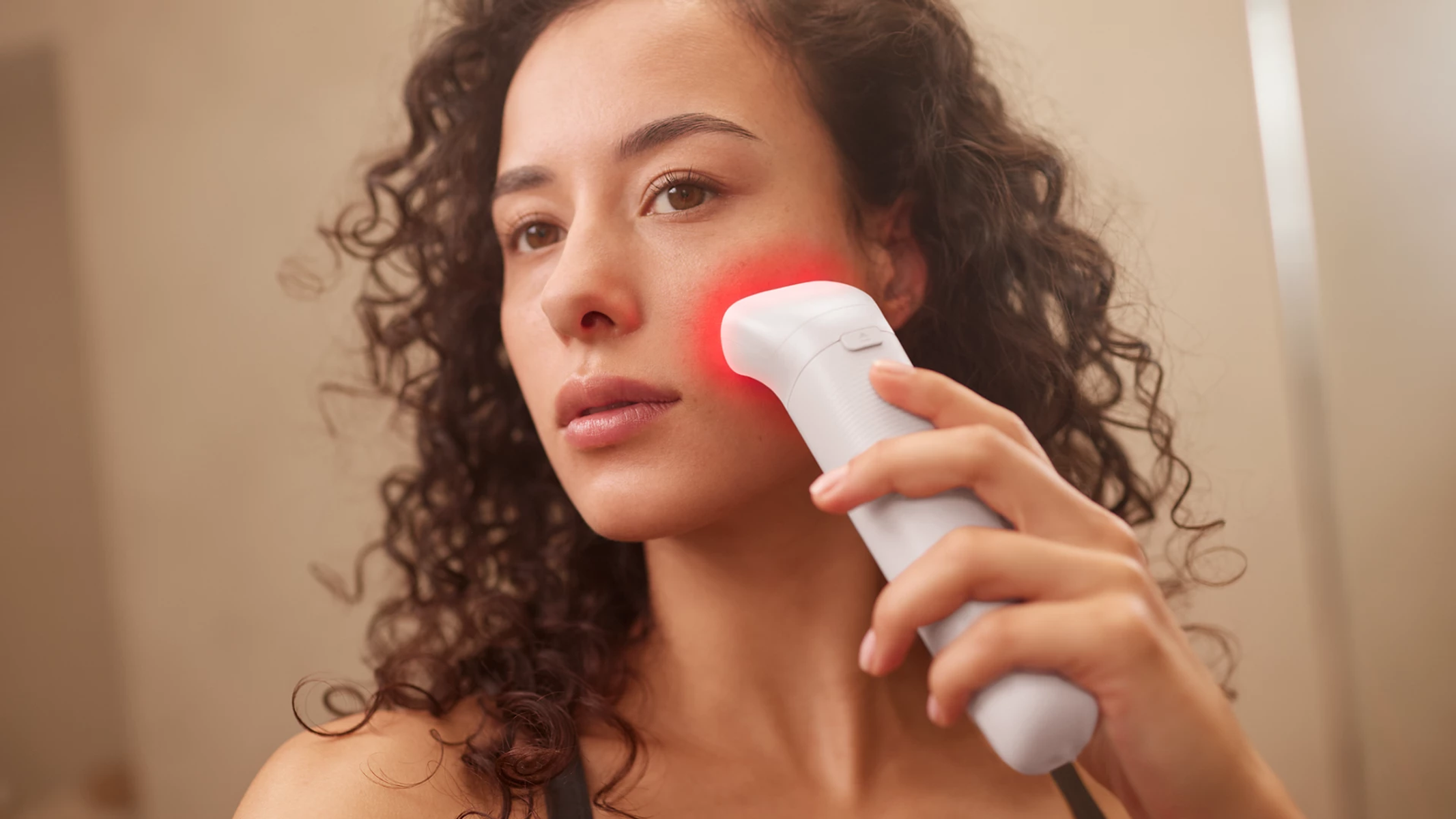 Una donna con capelli ricci usa un dispositivo di cura della pelle bianco con luce rossa sulla guancia.