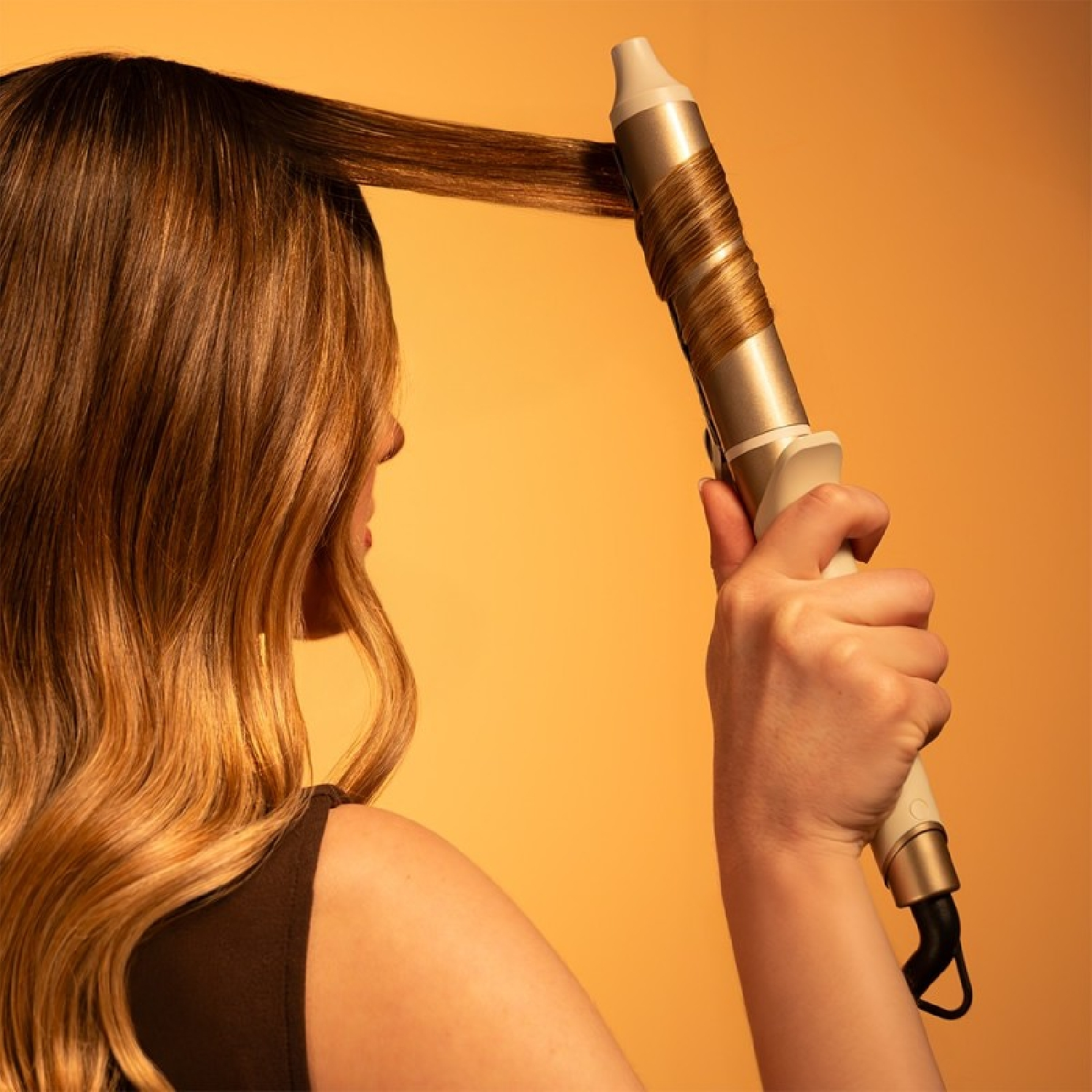 Una mujer se peina el pelo castaño con un rizador sobre fondo naranja.