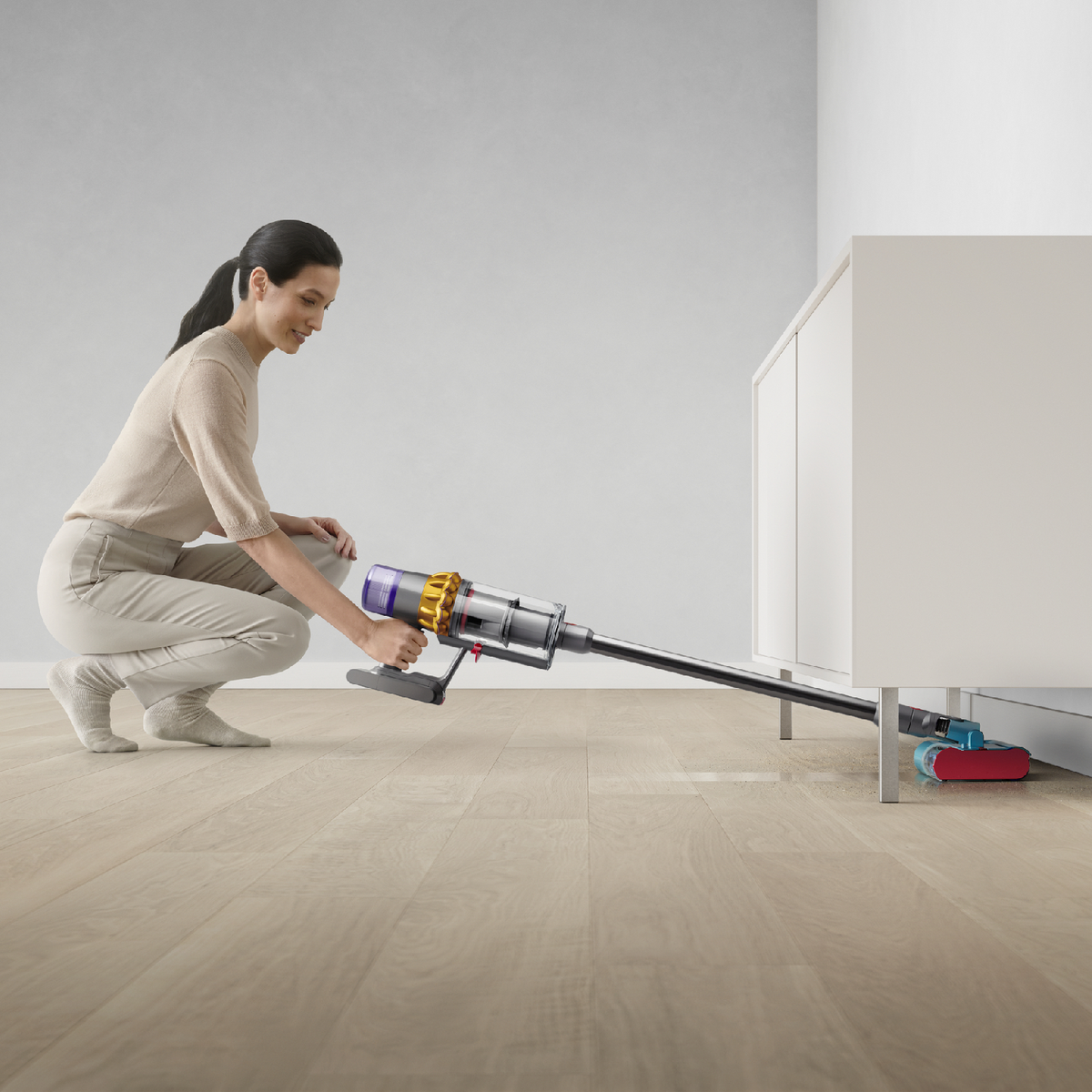Una donna si accovaccia su un pavimento in legno, usando un aspirapolvere senza fili sotto un mobile bianco.