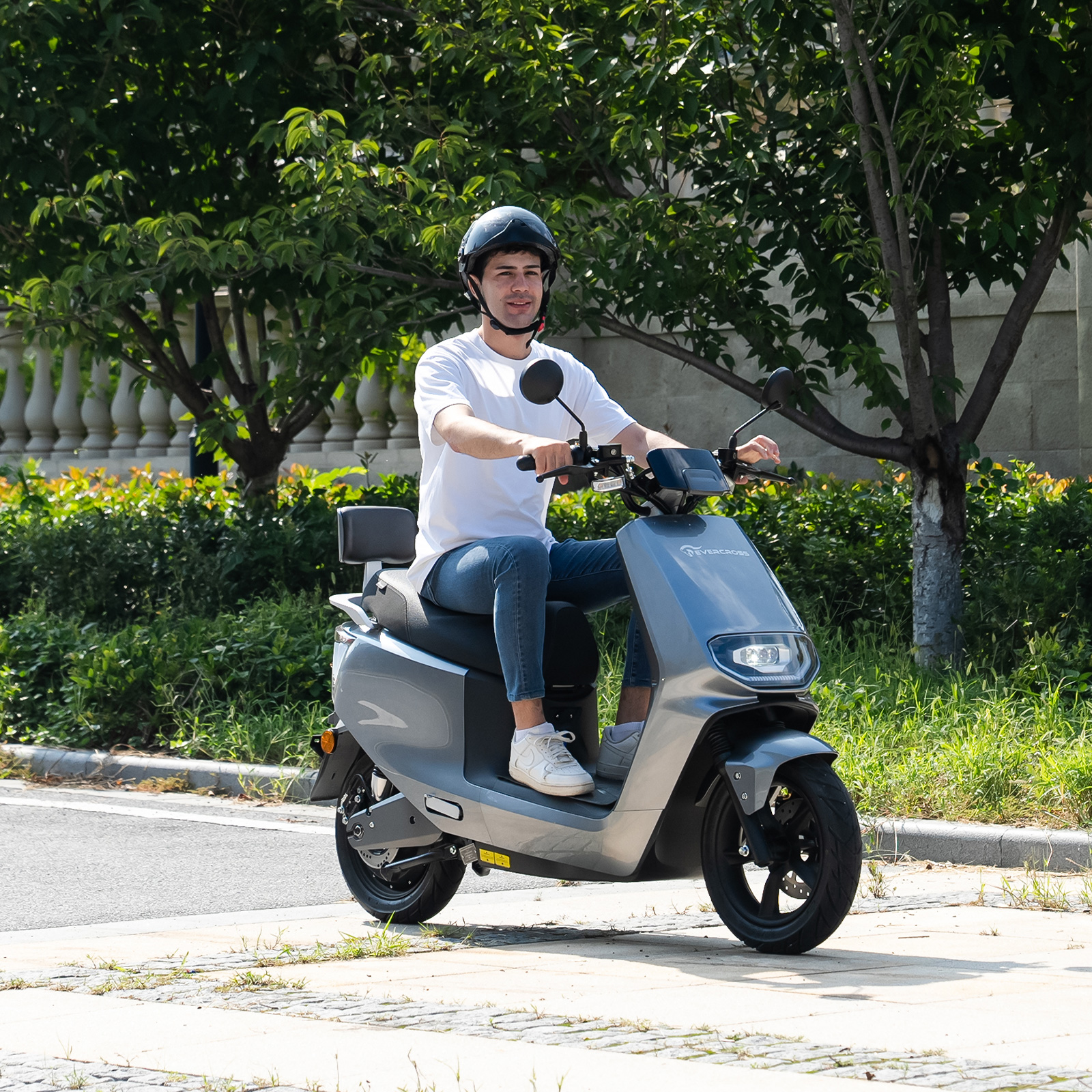 Ein Mann mit Helm fährt einen grauen Elektroroller auf einem Gehweg neben Bäumen.
