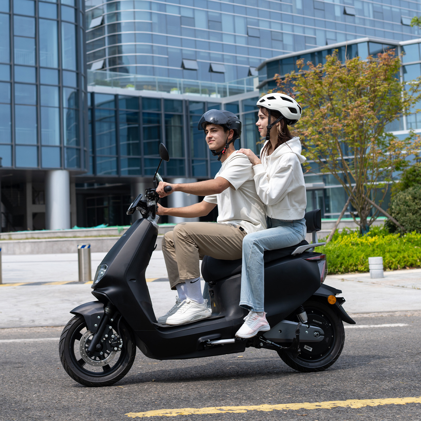 Ein Mann und eine Frau fahren auf einem schwarzen Roller vor einem modernen Gebäude.
