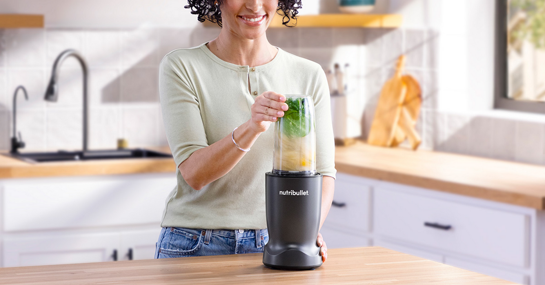 Frau mit Nutribullet Mixer mit grünen Blättern und Obst, in einer Küche stehend.