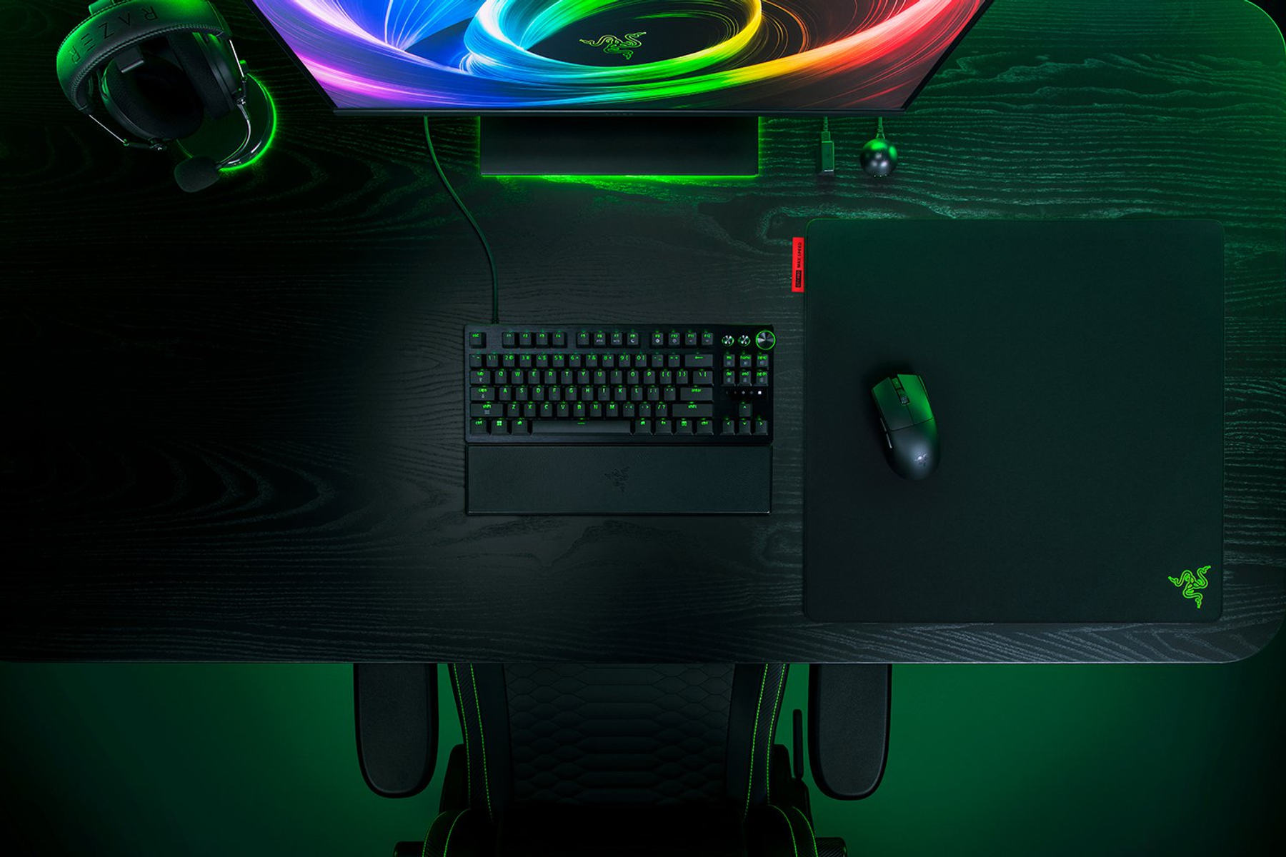 Un escritorio con teclado, ratón, auriculares y monitor en tonos oscuros con iluminación verde.