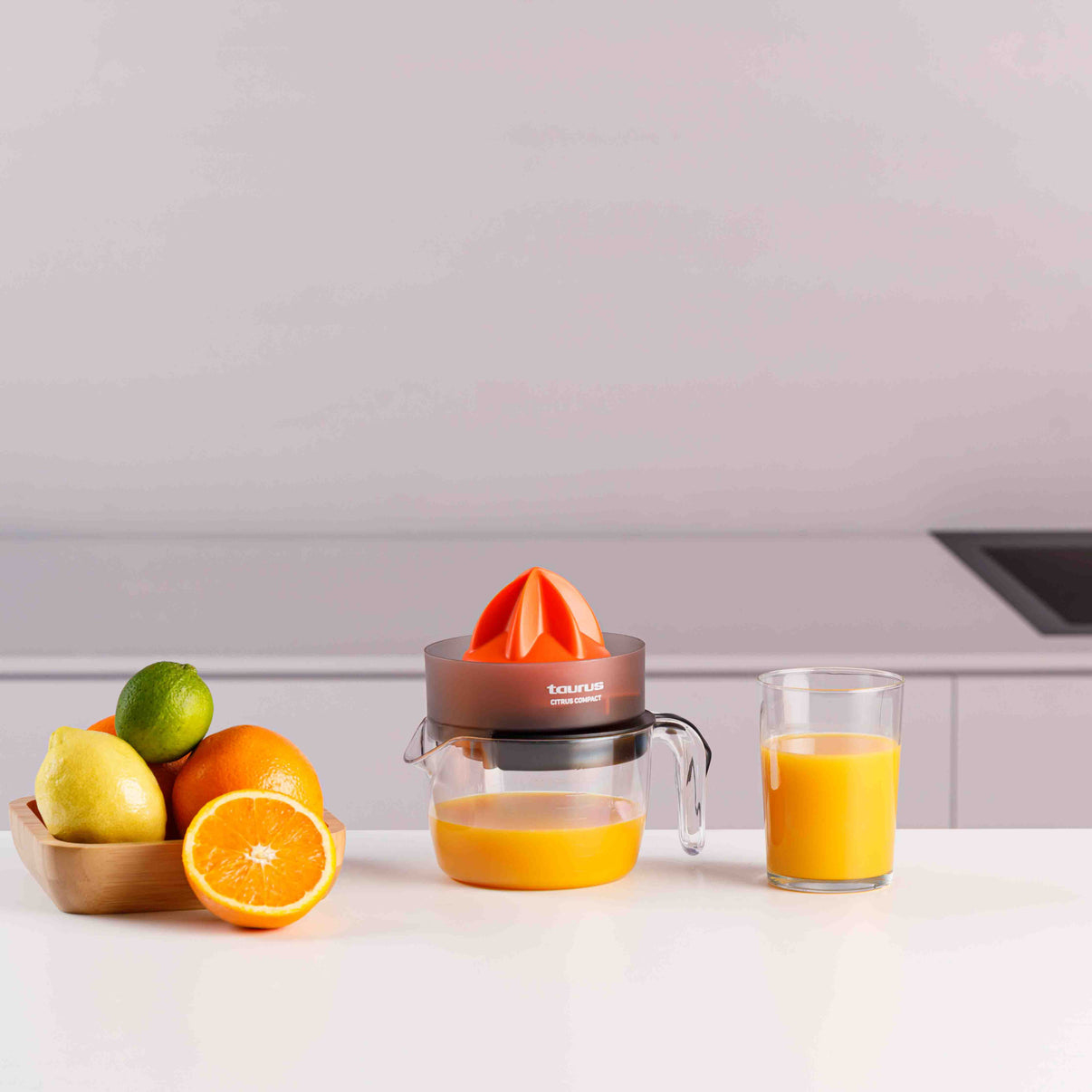 Exprimidor con naranjas, limón, lima y vaso de zumo en mostrador blanco.