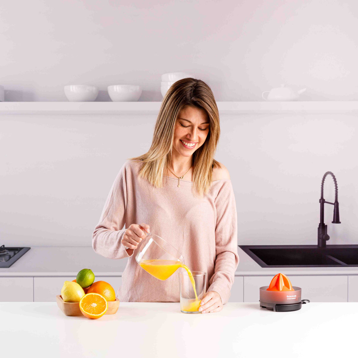 Una mujer vierte jugo de naranja de una jarra en un vaso en una cocina luminosa.