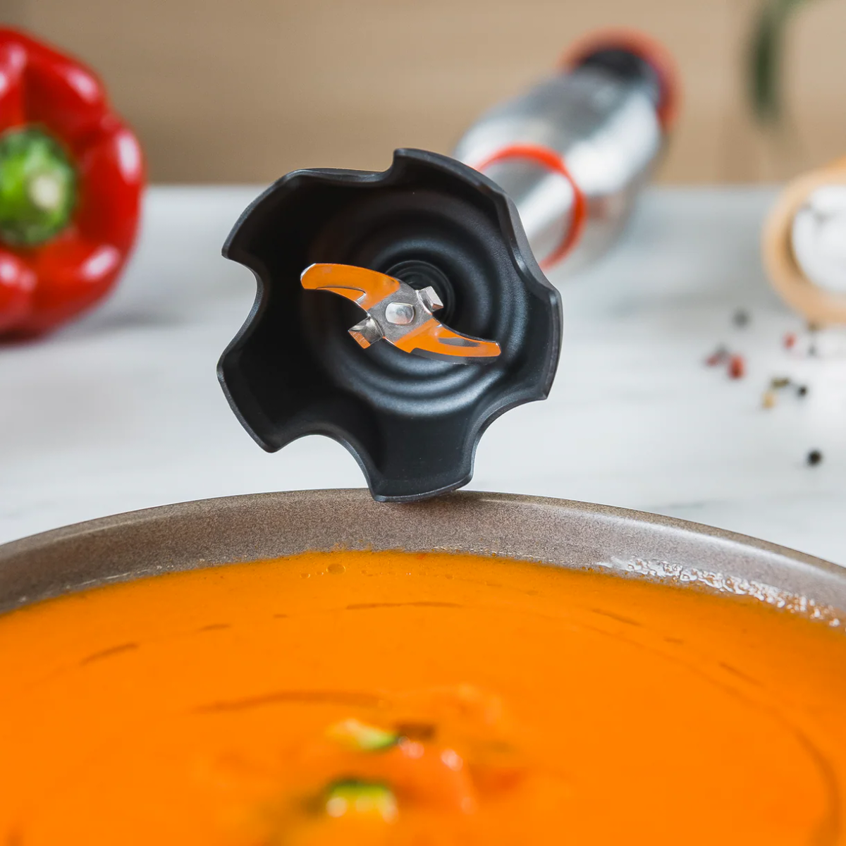 Lama di un frullatore a immersione su una ciotola di zuppa arancione, vicino a un peperone rosso.