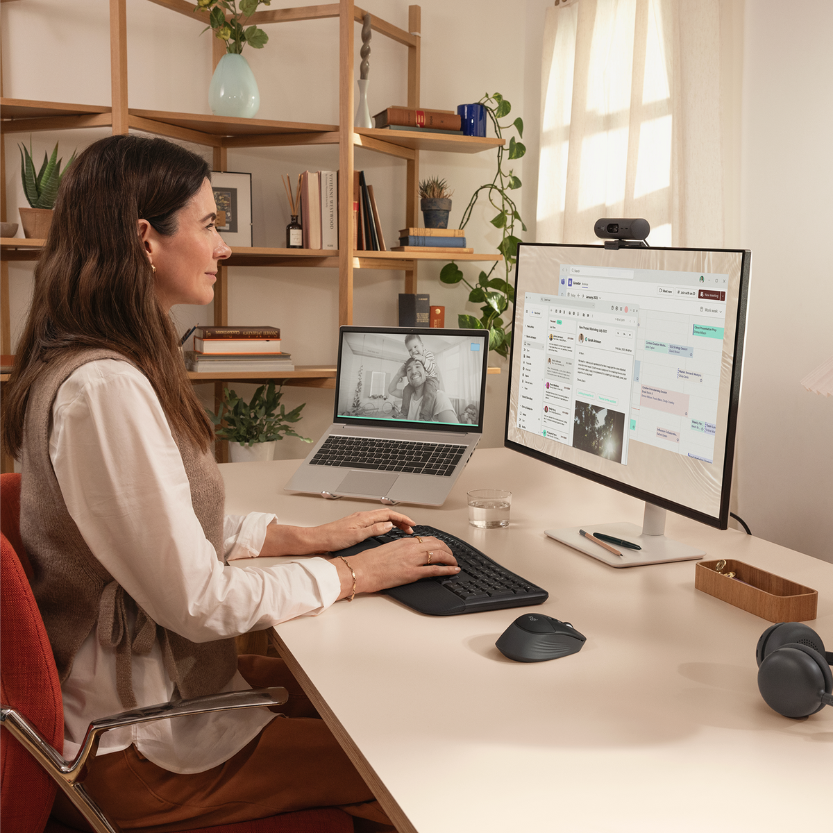 Donna alla scrivania, digita sulla tastiera. Laptop mostra foto famiglia. Monitor mostra lavoro.