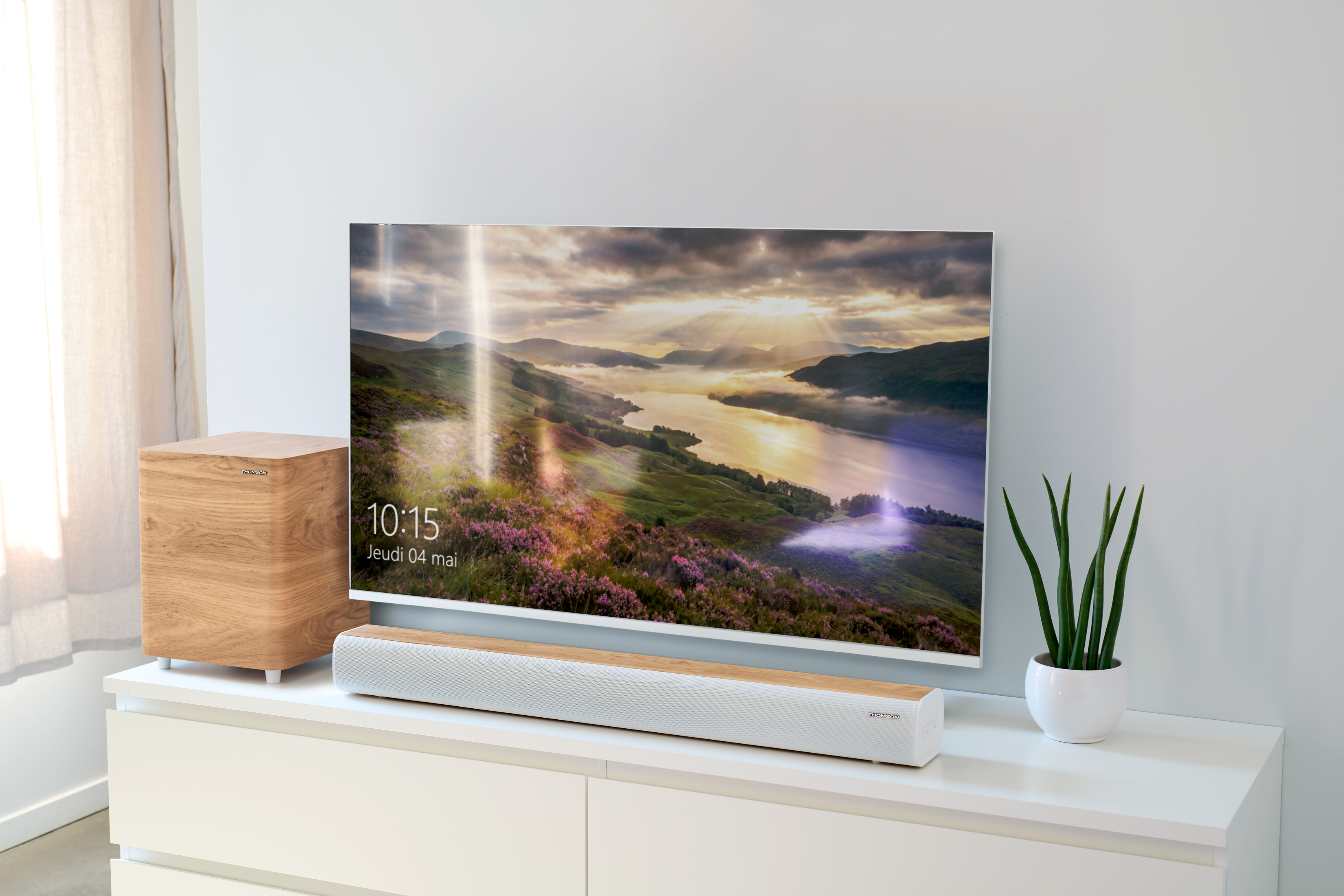 Subwoofer de madera, barra de sonido y TV con paisaje montañoso sobre un mueble blanco.