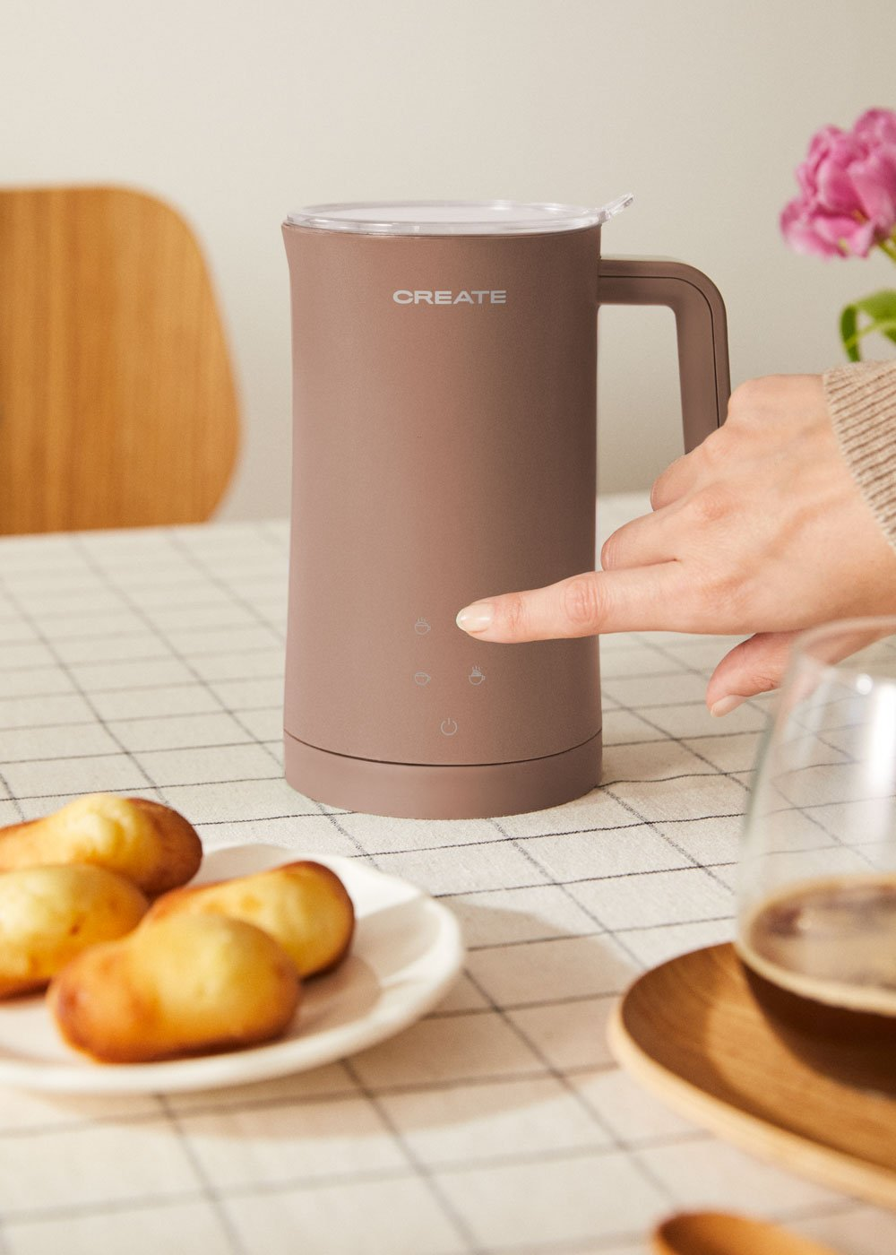Una persona presiona botones en un espumador de leche marrón junto a madeleines y café.