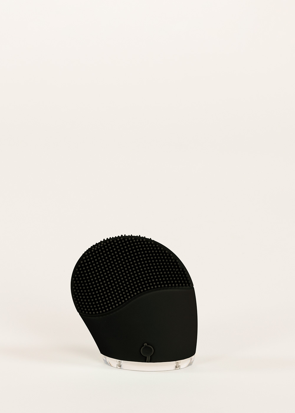 Un cepillo de limpieza facial negro con cerdas de silicona y base transparente descansa sobre superficie blanca.