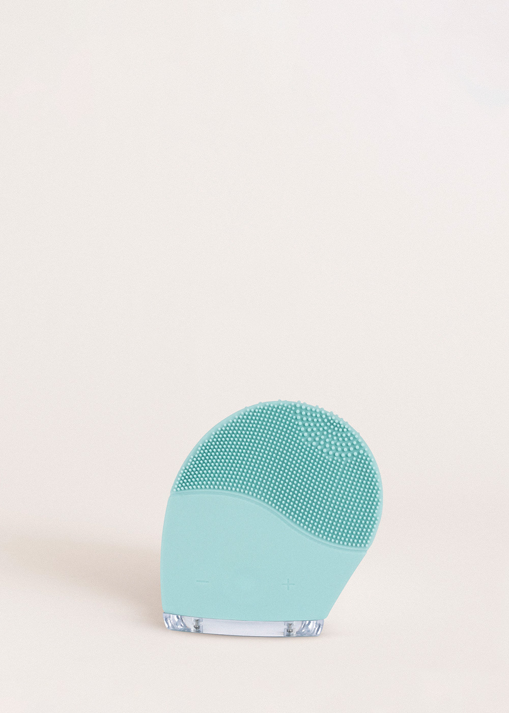 Un cepillo limpiador facial de silicona verde azulado con base transparente sobre fondo rosa claro.