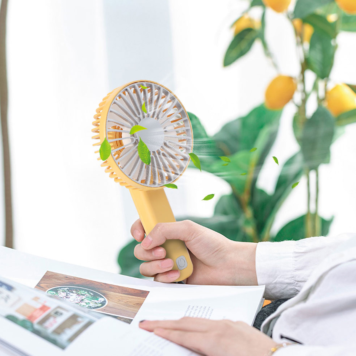 Eine Hand hält einen gelben Ventilator, der Blätter wirbelt. Im Hintergrund sind Zitronen und Blätter.
