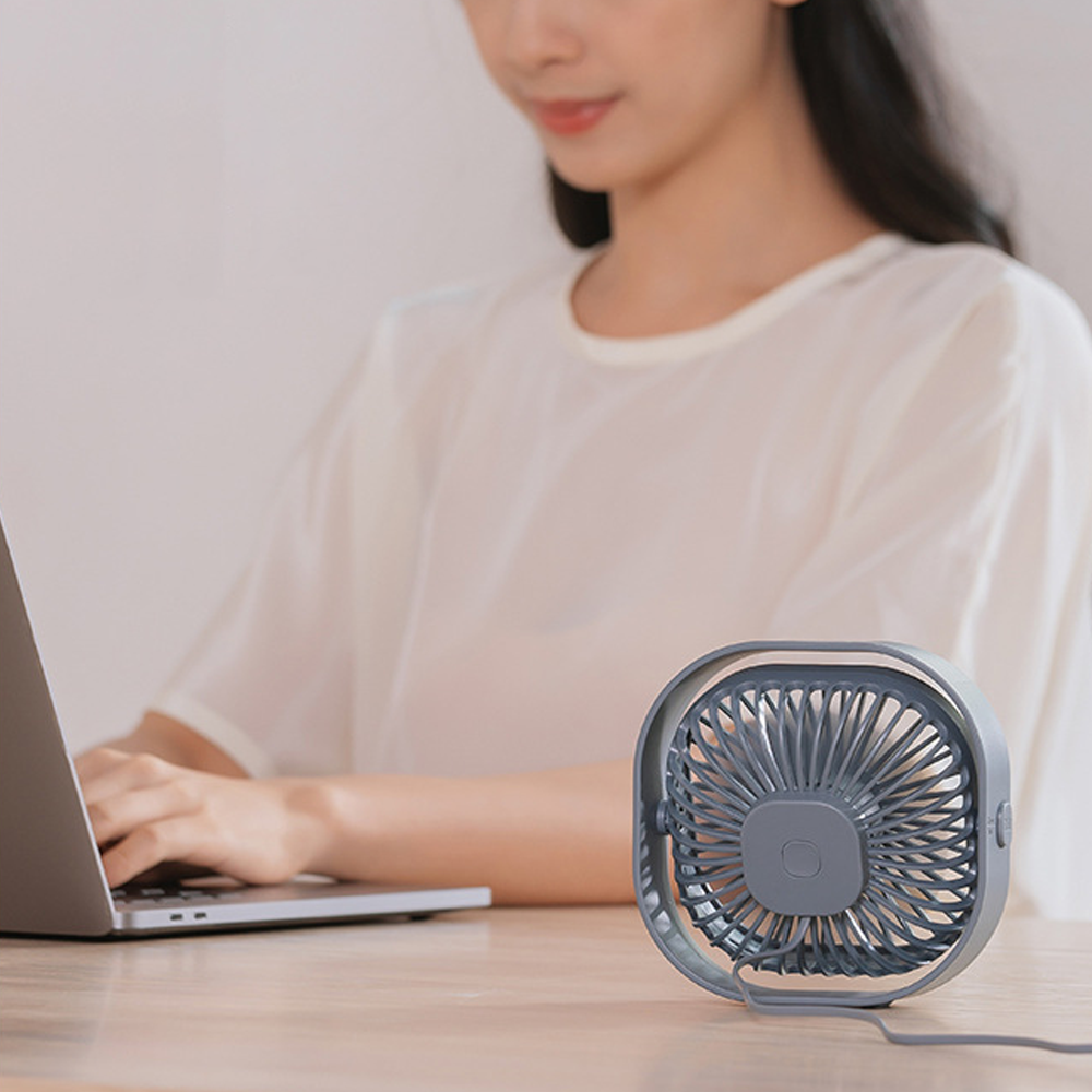 Eine Frau tippt auf einem Laptop, ein kleiner grauer Ventilator steht daneben.