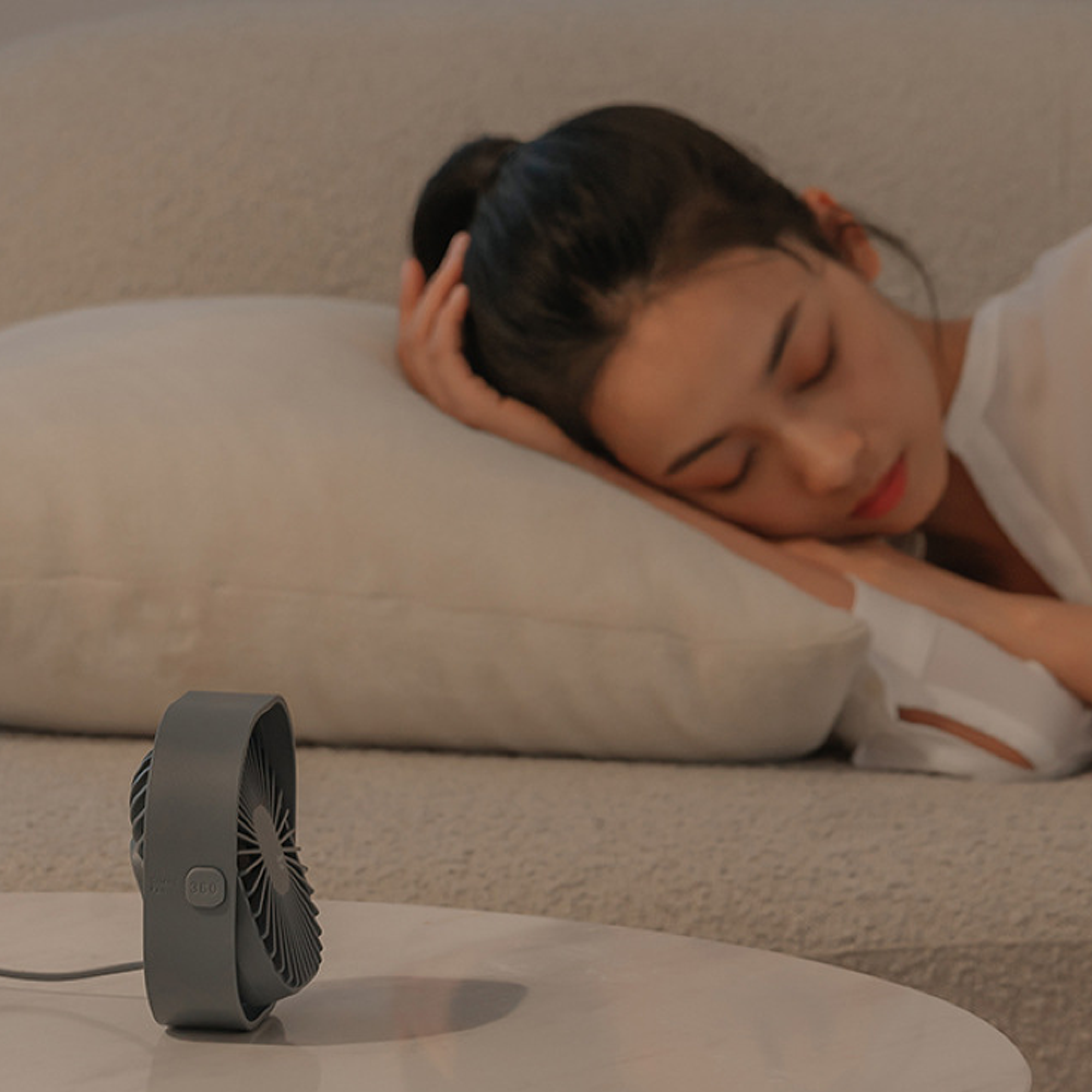 Ein grauer Ventilator steht auf einem Tisch, während eine Frau auf einem Sofa schläft.