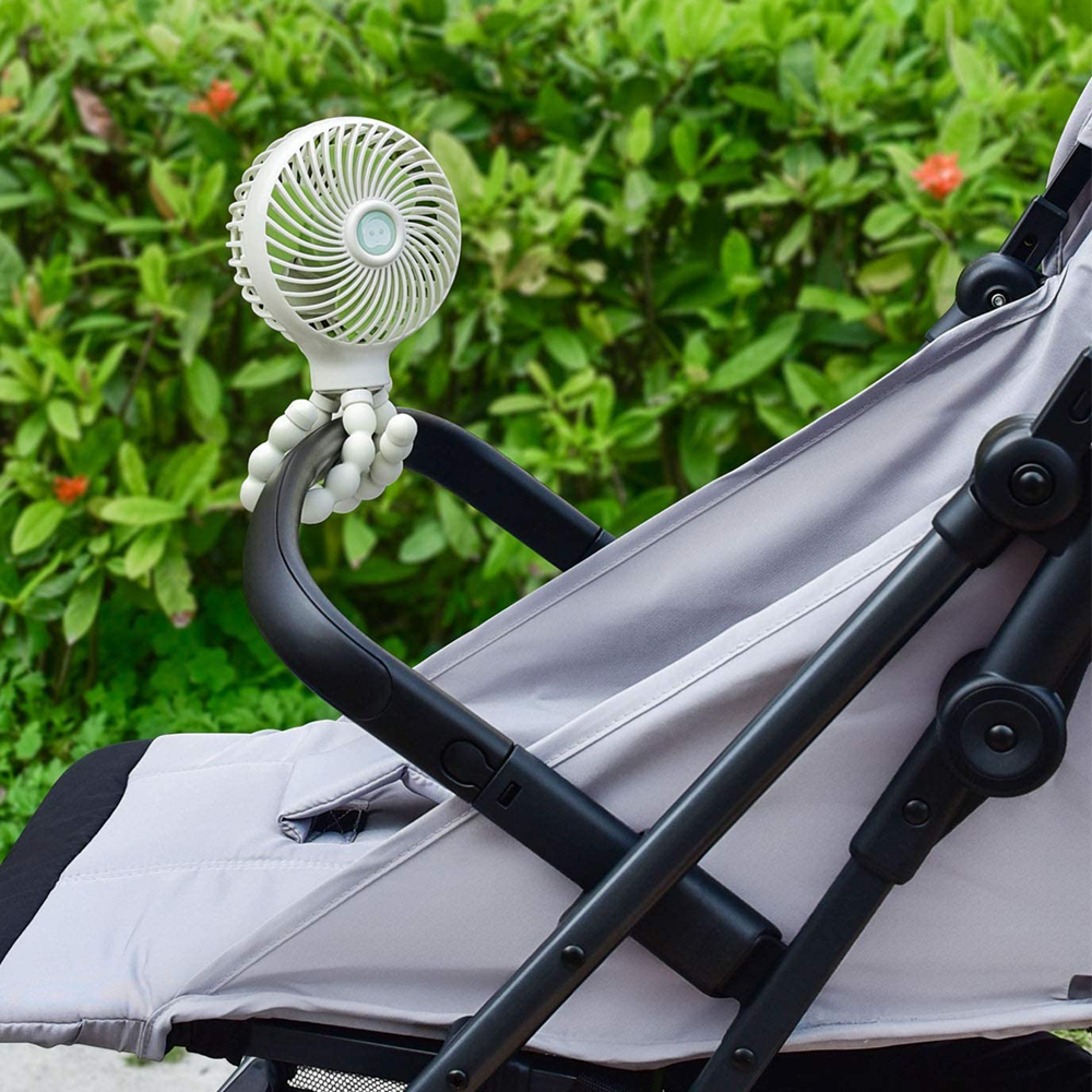 Ein weißer Ventilator ist an einem Kinderwagen befestigt. Im Hintergrund ist grünes Laub.