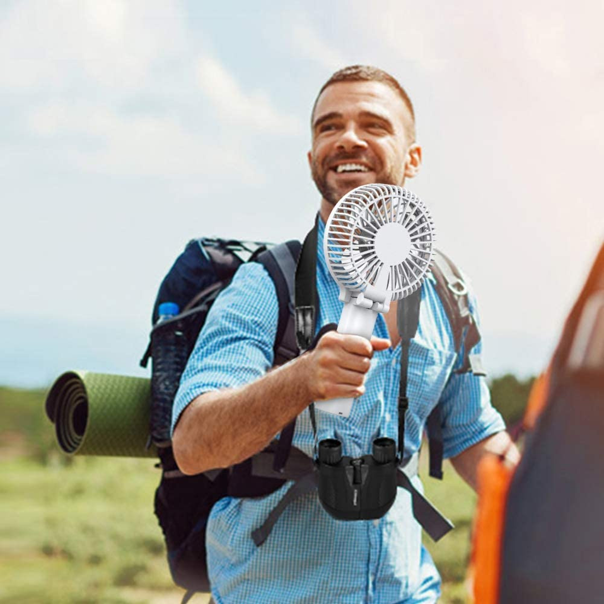 Ein Mann mit Rucksack hält einen kleinen Ventilator, Fernglas um den Hals, draußen.