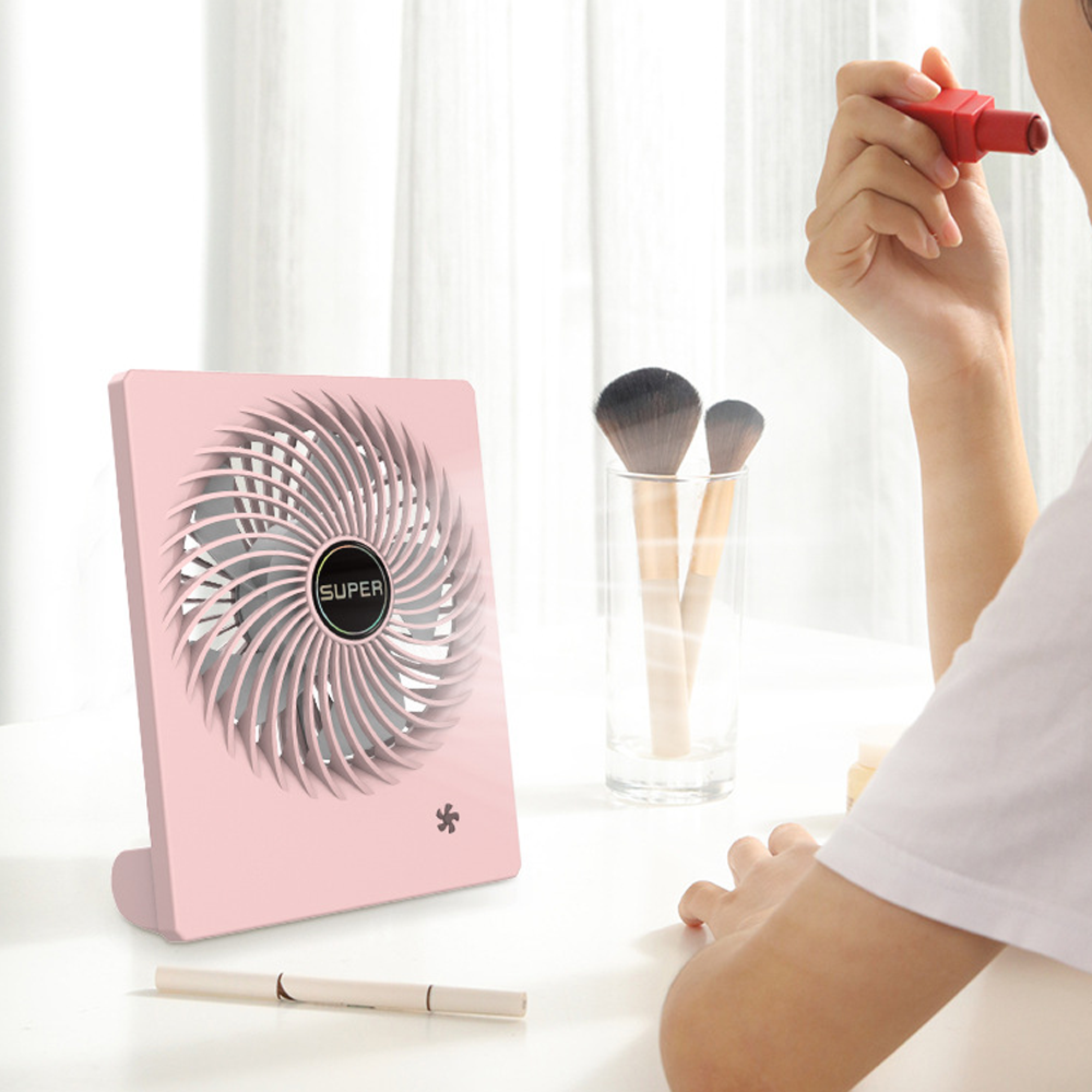 Ein rosa Ventilator steht auf einem Tisch mit Kosmetikpinseln und einer Person, die Lippenstift hält.