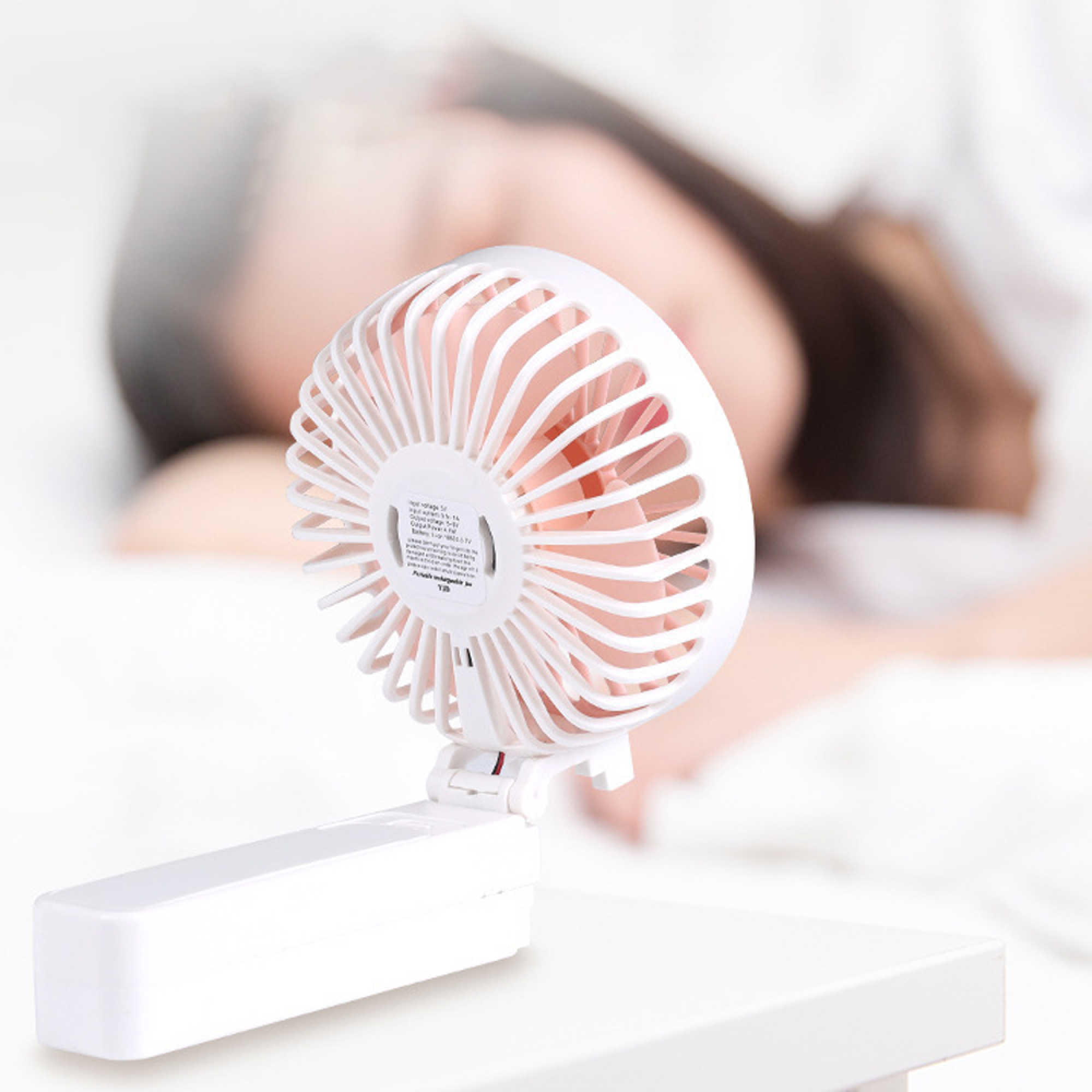 Ein kleiner weißer Ventilator steht auf einem Tisch vor einer schlafenden Person.