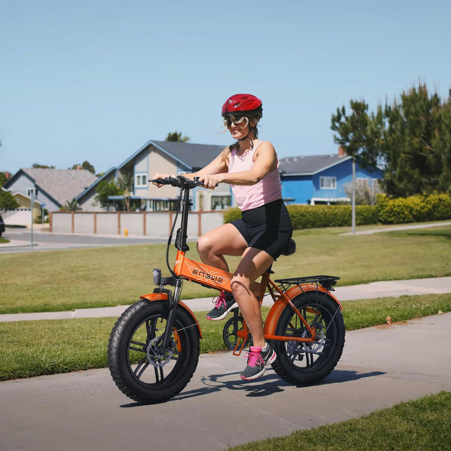Una mujer con casco rojo monta una bicicleta plegable naranja en una acera con casas.