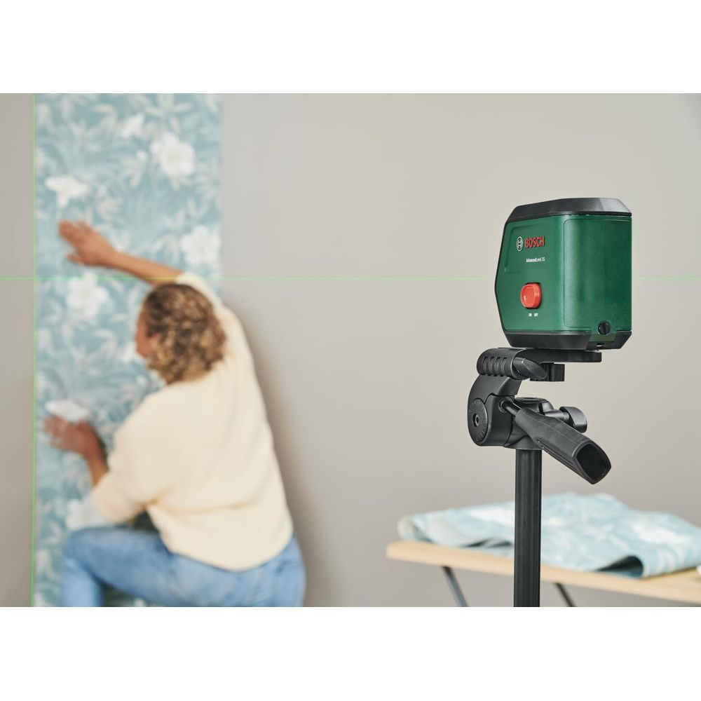 Grüner Laser Nivellierer auf Stativ neben Person, die Tapete anbringt.