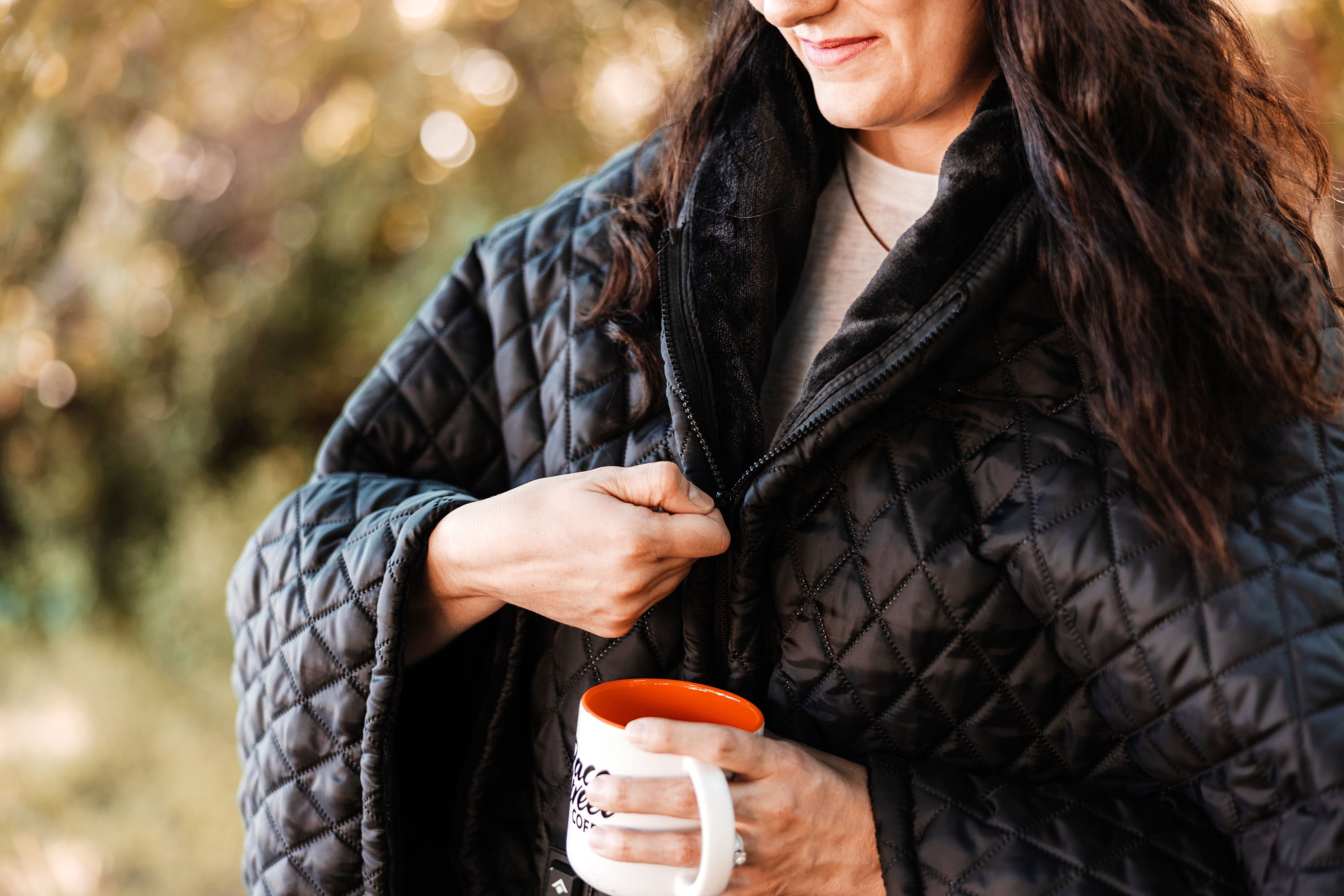 Eine Frau mit dunklem Haar schließt eine gesteppte schwarze Jacke und hält eine weiße Tasse.