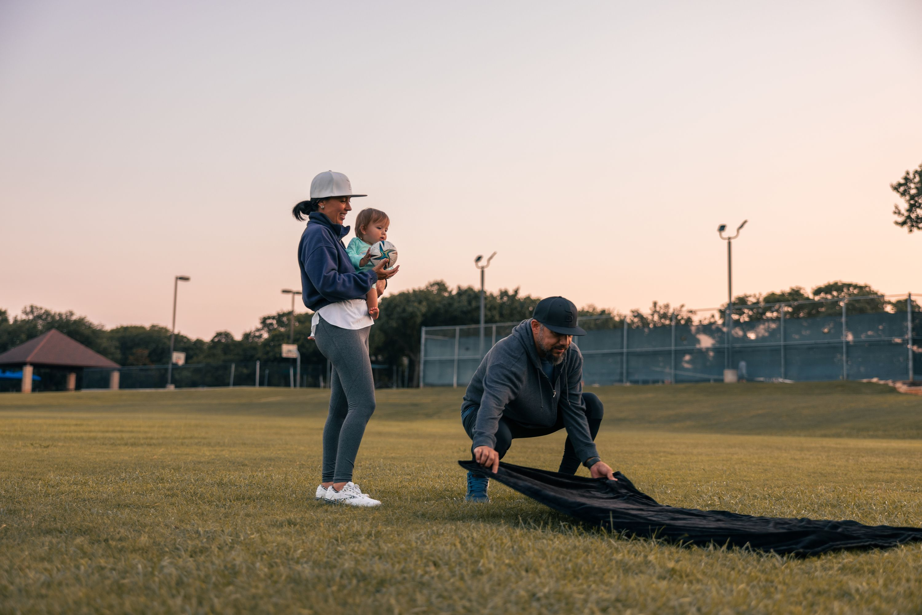 Ein Mann legt eine schwarze Decke auf Gras, eine Frau hält ein Baby, ein Park im Hintergrund.
