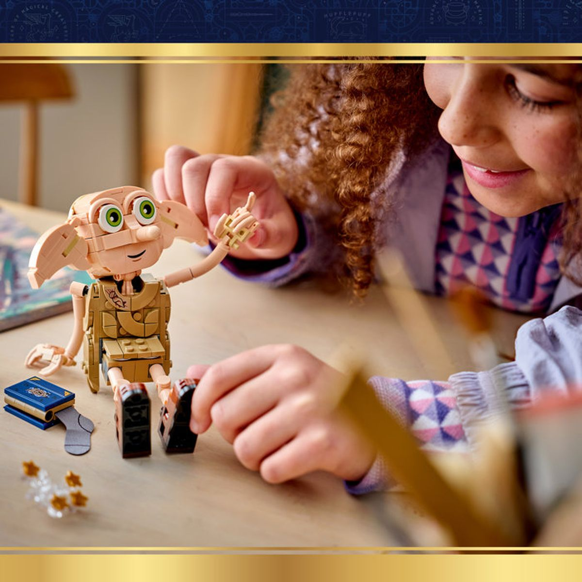Dziewczynka bawi się figurką Lego Dobby, która siedzi przy drewnianym stole obok niebieskiej książki.