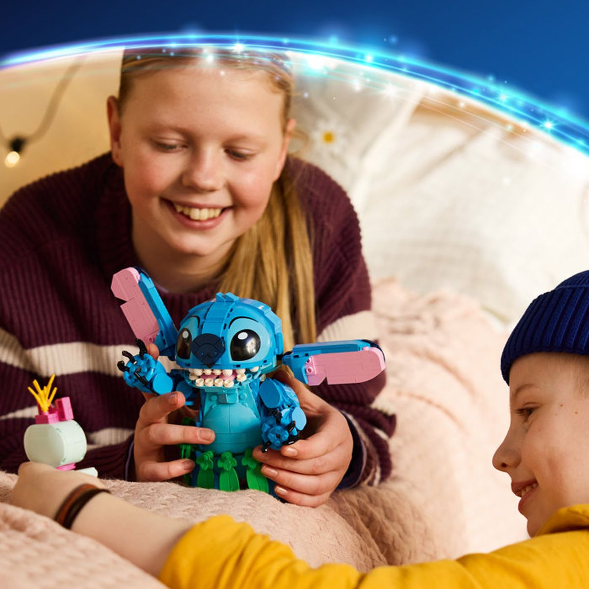 Dwoje dzieci trzyma niebieskiego Stitcha z Lego z różowymi skrzydłami i zielonymi nogami.