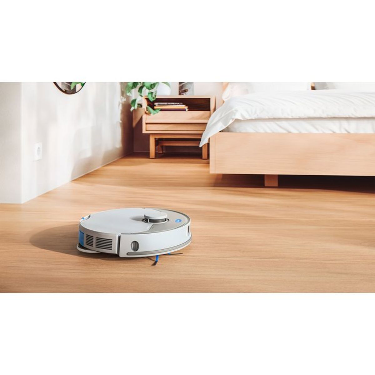 Een witte robotstofzuiger op een lichte houten vloer bij een bed.