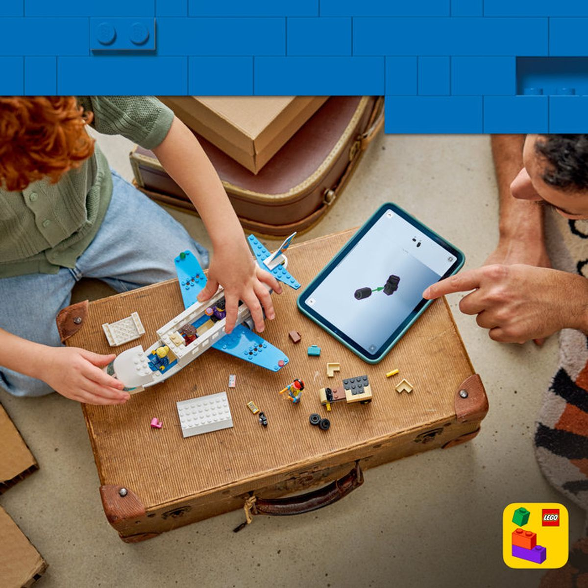Çocuk, yetişkin tabletle yönlendirirken bavul üzerinde mavi Lego uçak yapıyor.