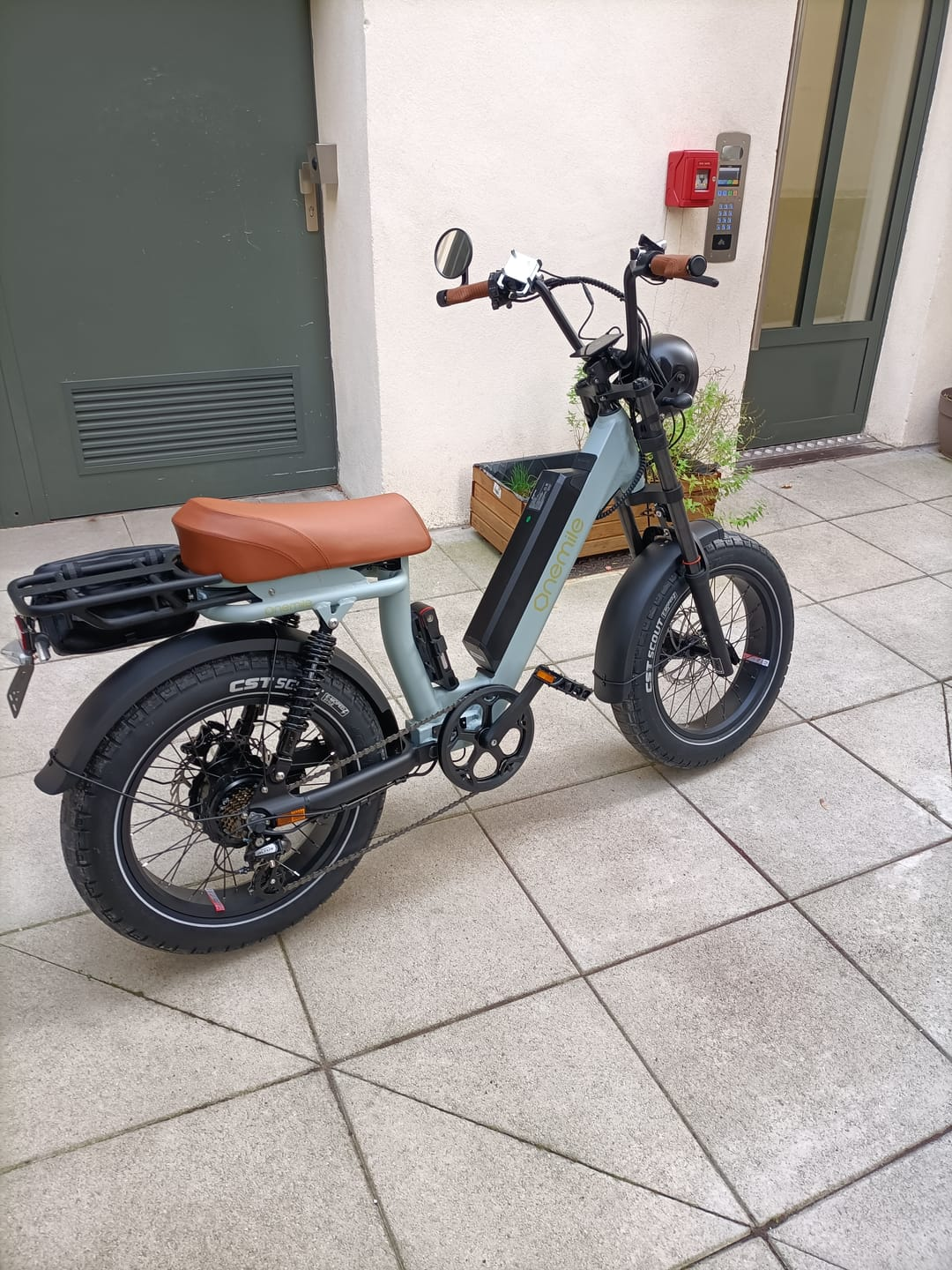 Una e-bike grigio chiaro con sella e portapacchi marrone si trova su un patio piastrellato.