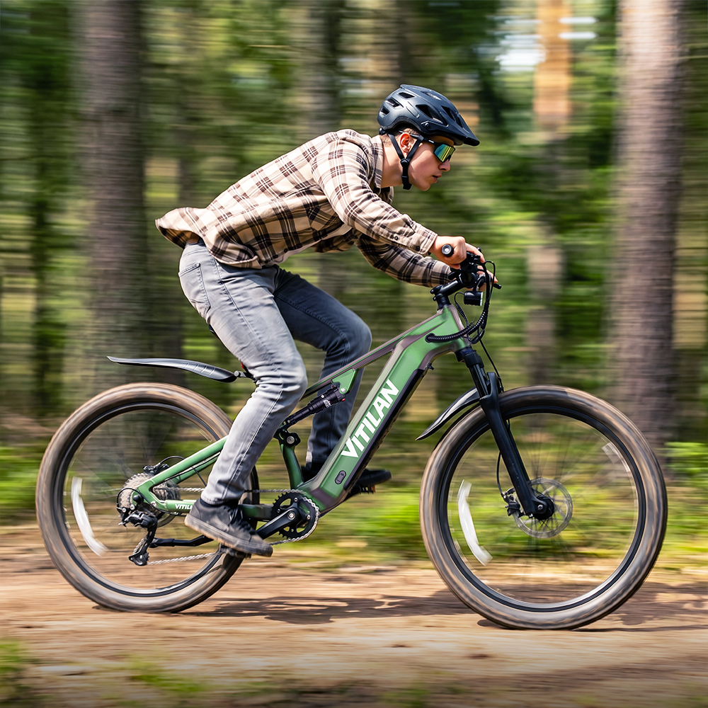 Un hombre monta una e-bike verde por un sendero forestal. Lleva casco, gafas y camisa a cuadros.