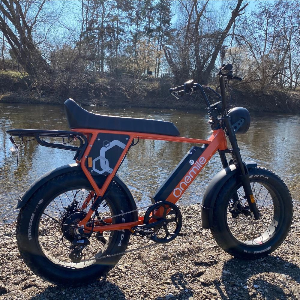 Una e-bike arancione riposa su una riva ghiaiosa accanto a un fiume con alberi sullo sfondo.