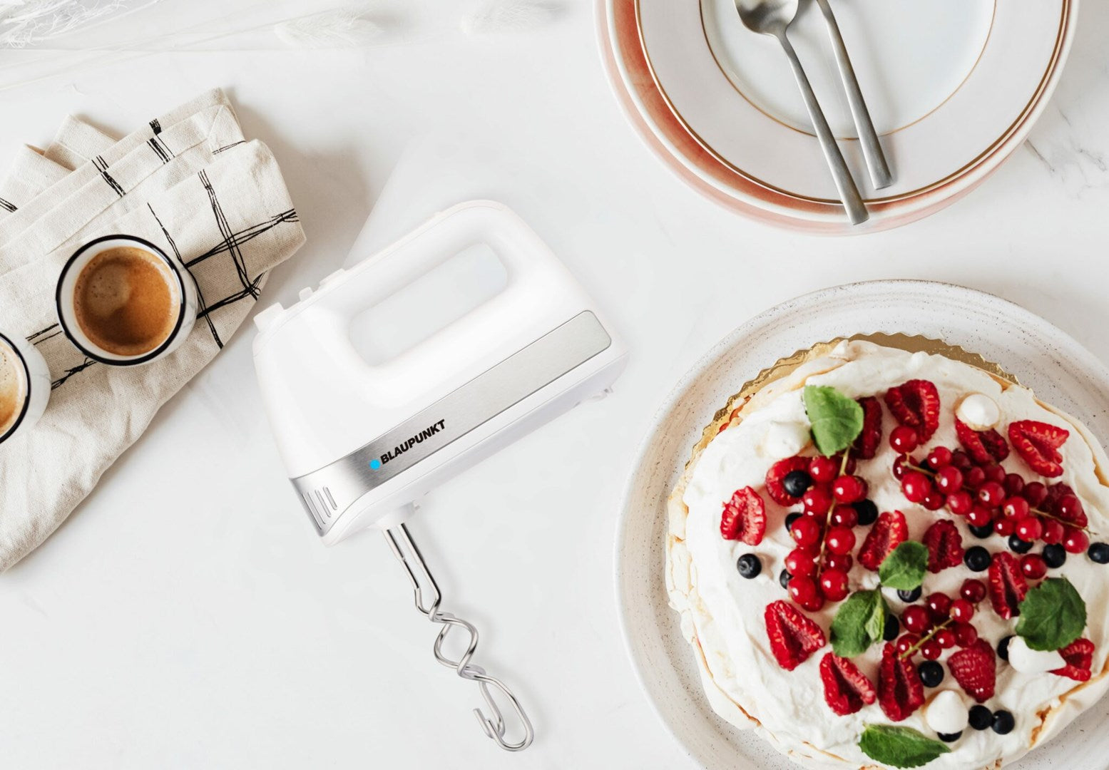 Ein weißer Handmixer neben einer Pavlova-Torte mit Beeren und zwei Tassen Kaffee.