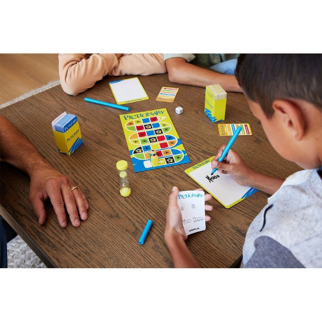Bambini giocano a Pictionary su tavolo di legno con tabellone, carte e penne.