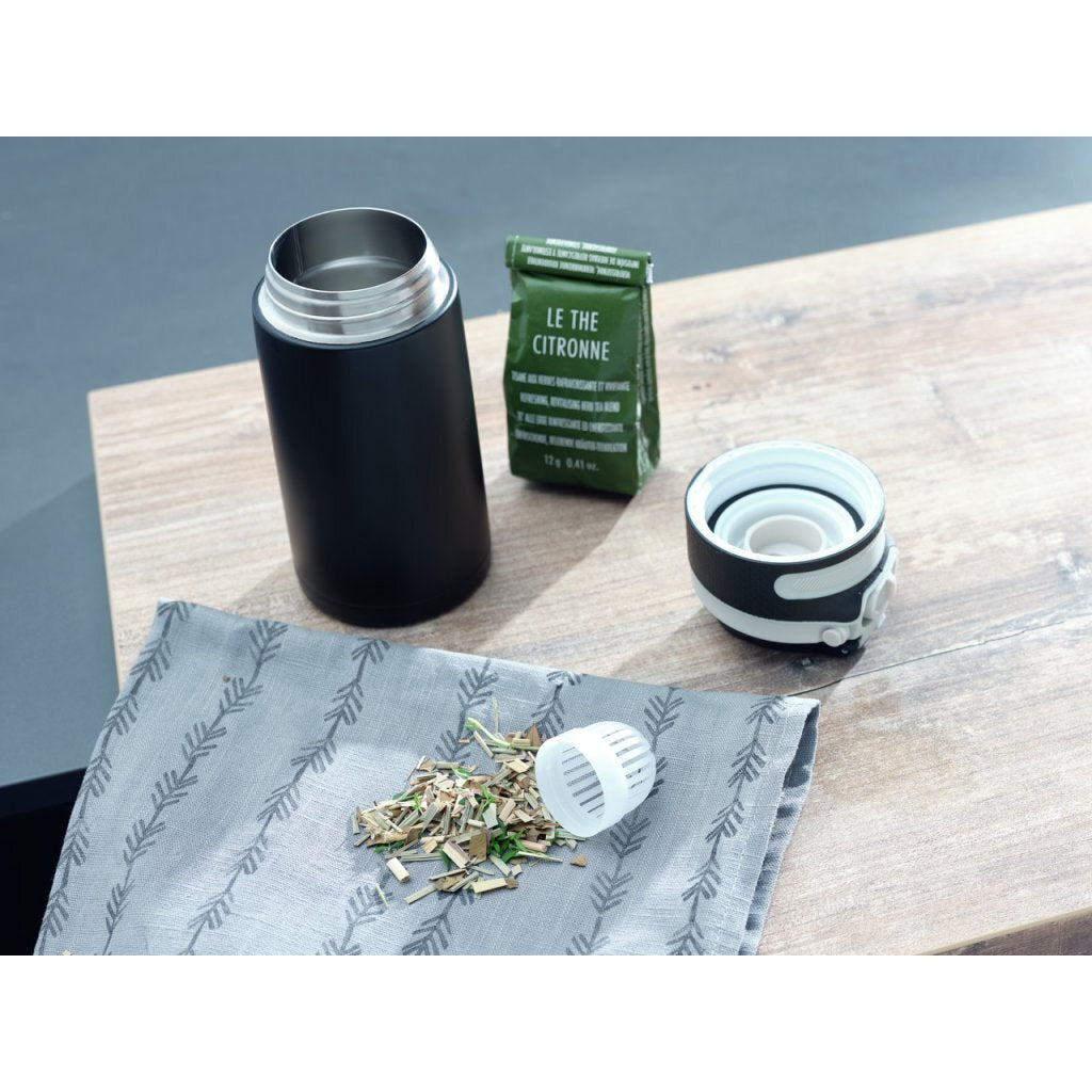 Schwarze Thermoskanne, Teepackung, Deckel und Kräuter auf Holztisch.