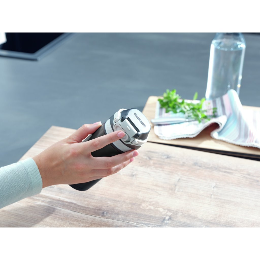 Hand hält schwarze Trinkflasche mit weißem Verschluss. Tisch mit Tuch, Kräutern und Wasserflasche.