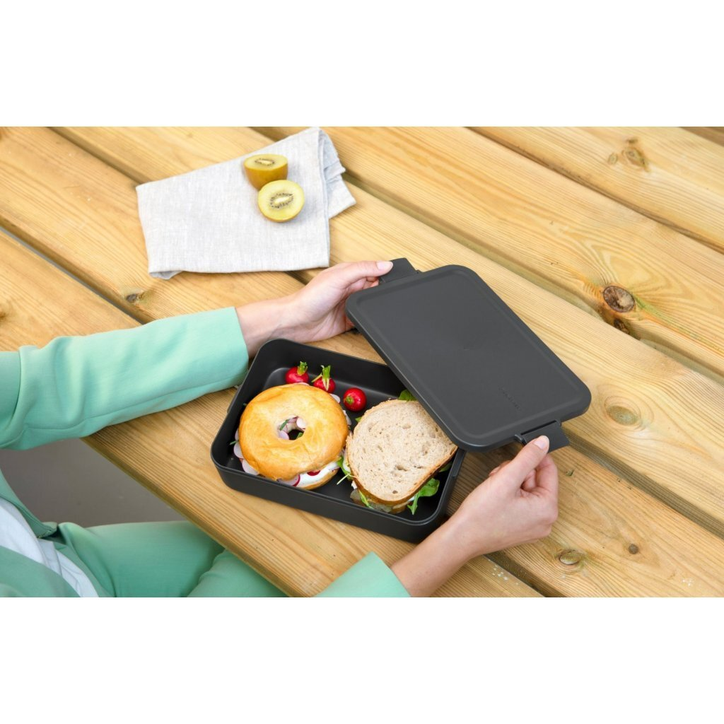 Persona che chiude un portapranzo con bagel, sandwich e ravanelli su tavolo in legno.