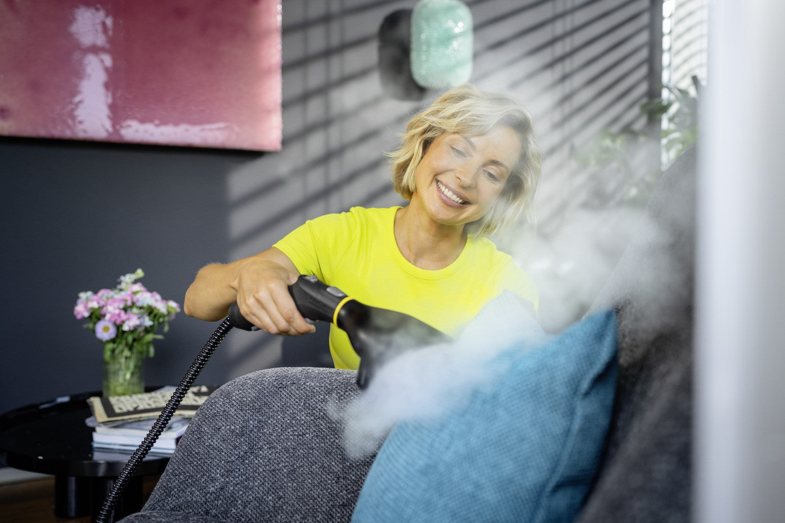 Eine Frau reinigt mit einem Dampfreiniger ein Sofa. Sie lächelt.